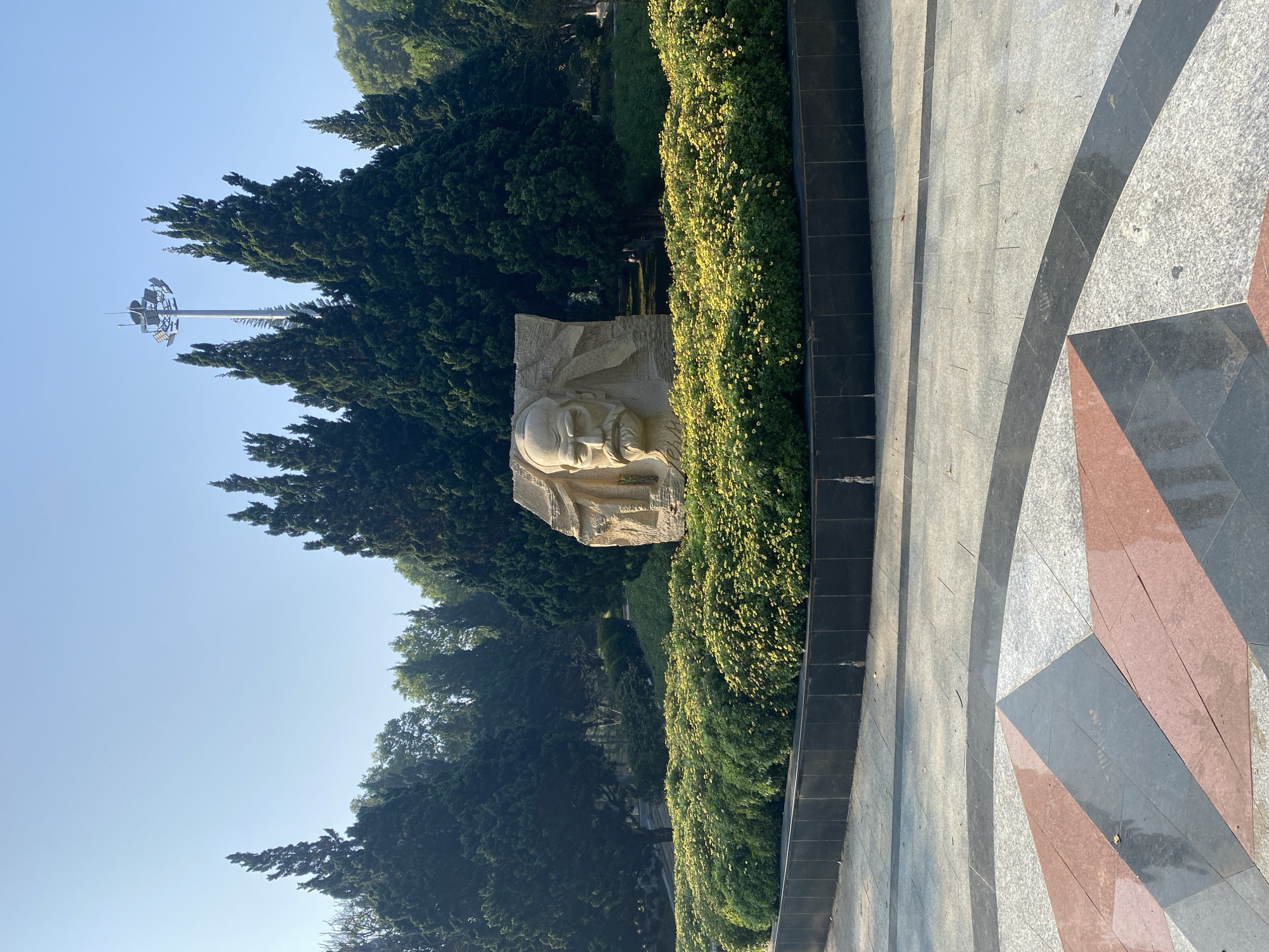 The image shows a stone bust monument of Dr. Alexandre Yersin in Da Lat, Vietnam. The bust, carved in light-colored stone, is set within a lush, green landscape, bordered by flowering shrubs. Tall evergreen trees form a backdrop to the monument. A modern light pole is visible in the background. To the right, a decorative paved area with geometric patterns in gray, black, and red is visible. The sky is clear and light blue.