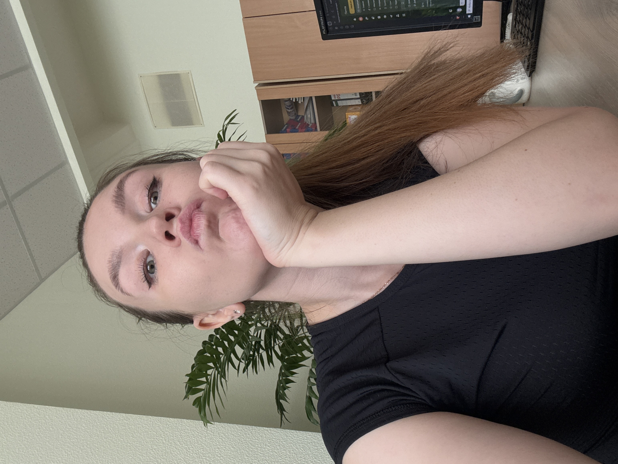 A young woman is shown from the chest up in a close-up selfie. She has her right hand resting on her chin, her lips pursed. Her eyes are open and looking forward. She is wearing a black t-shirt. To her left is a large green plant. Behind her, on the right, is a wooden cabinet with shelves and a computer monitor displaying a screen with Russian text. The scene appears to be indoors, possibly an office or home environment. The lighting is bright and natural, suggesting daytime. There is no indication of the location being in Izhevsk, Russia.