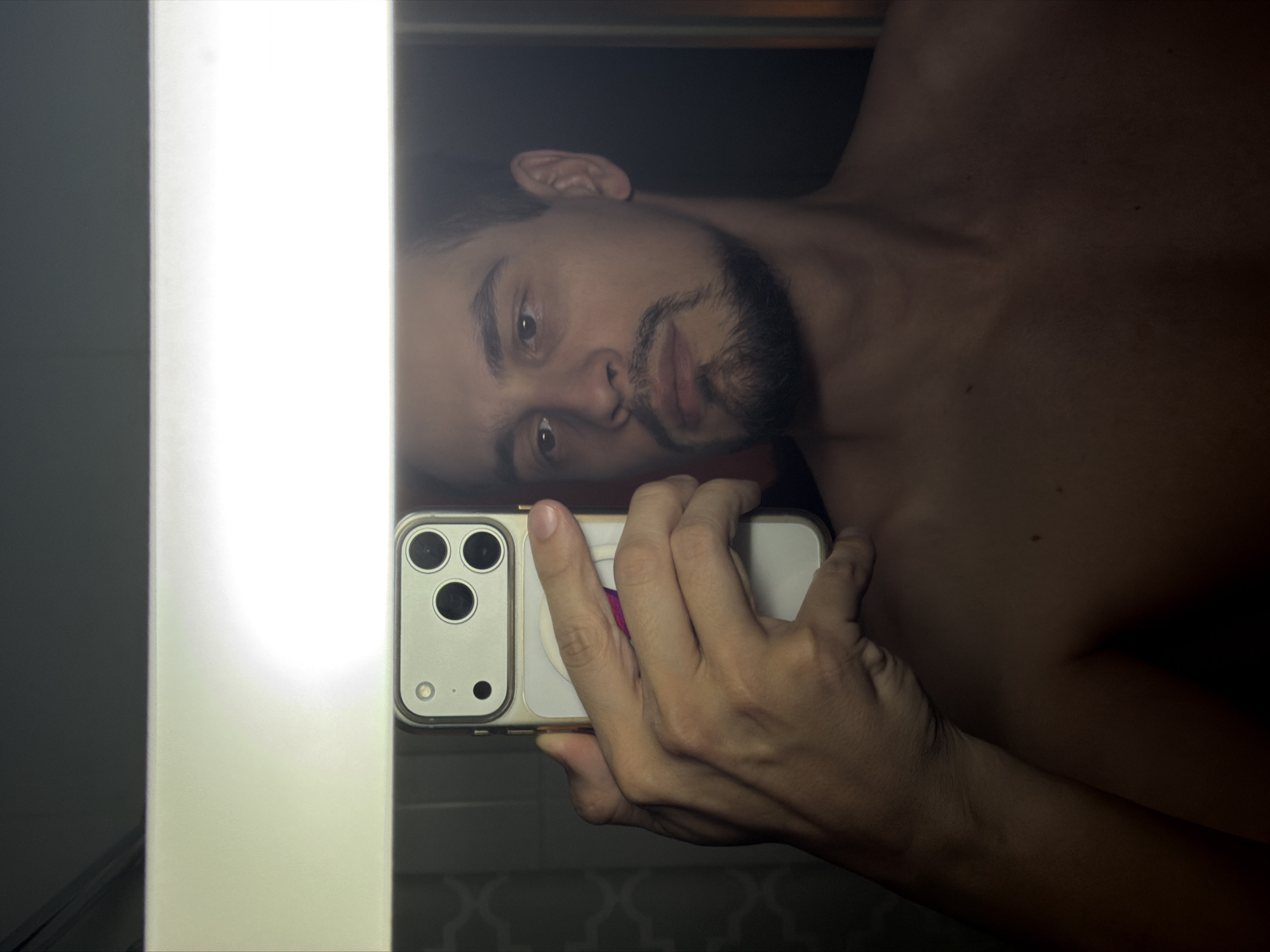 A man is taking a selfie in what appears to be a bathroom. He is holding a smartphone, possibly an iPhone, with three rear-facing cameras. He has facial hair and is shirtless. A bright, vertical light source illuminates one side of the image. The location in Krasnoyarsk, Russia, is not discernible from the image.