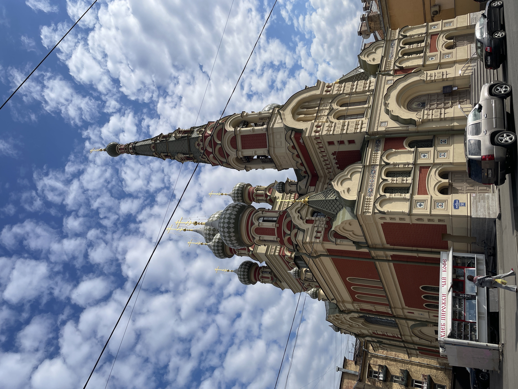 This image captures a vibrant street scene in Saint Petersburg, Russia, centered around a strikingly ornate church building under a bright, cloudy sky. The church, a prime example of Russian Orthodox architecture, features multiple onion domes, some with a checkered pattern and others gray, all topped with golden Orthodox crosses. A very tall, slender spire also culminates in a golden cross, dominating the upper portion of the structure. The building's facade is a rich blend of red brick and cream-colored plasterwork, adorned with intricate decorative elements, arched windows, and patterned tiles.
In the foreground, a city street is visible with two cars parked: a silver SUV and a black sedan. On the sidewalk, a white kiosk with red trim stands prominently. Text visible on the kiosk reads "ХЛЕБ ПИРОЖКИ ЧАЙ КОФЕ" (BREAD PIROGUES TEA COFFEE), indicating its offerings. A person, dressed in a jacket and scarf, is standing near the kiosk, suggesting typical daily activity. Above the kiosk, a blue street sign clearly displays "БОРОВАЯ" (Borovaya) and the number "52," identifying the street address.
The weather appears pleasant, with a clear blue sky interspersed with fluffy white clouds, indicating daytime. Overhead, dark power lines stretch across the frame, adding an urban element to the scene. The overall impression is one of a busy yet architecturally rich city environment, with the church standing as a magnificent landmark.