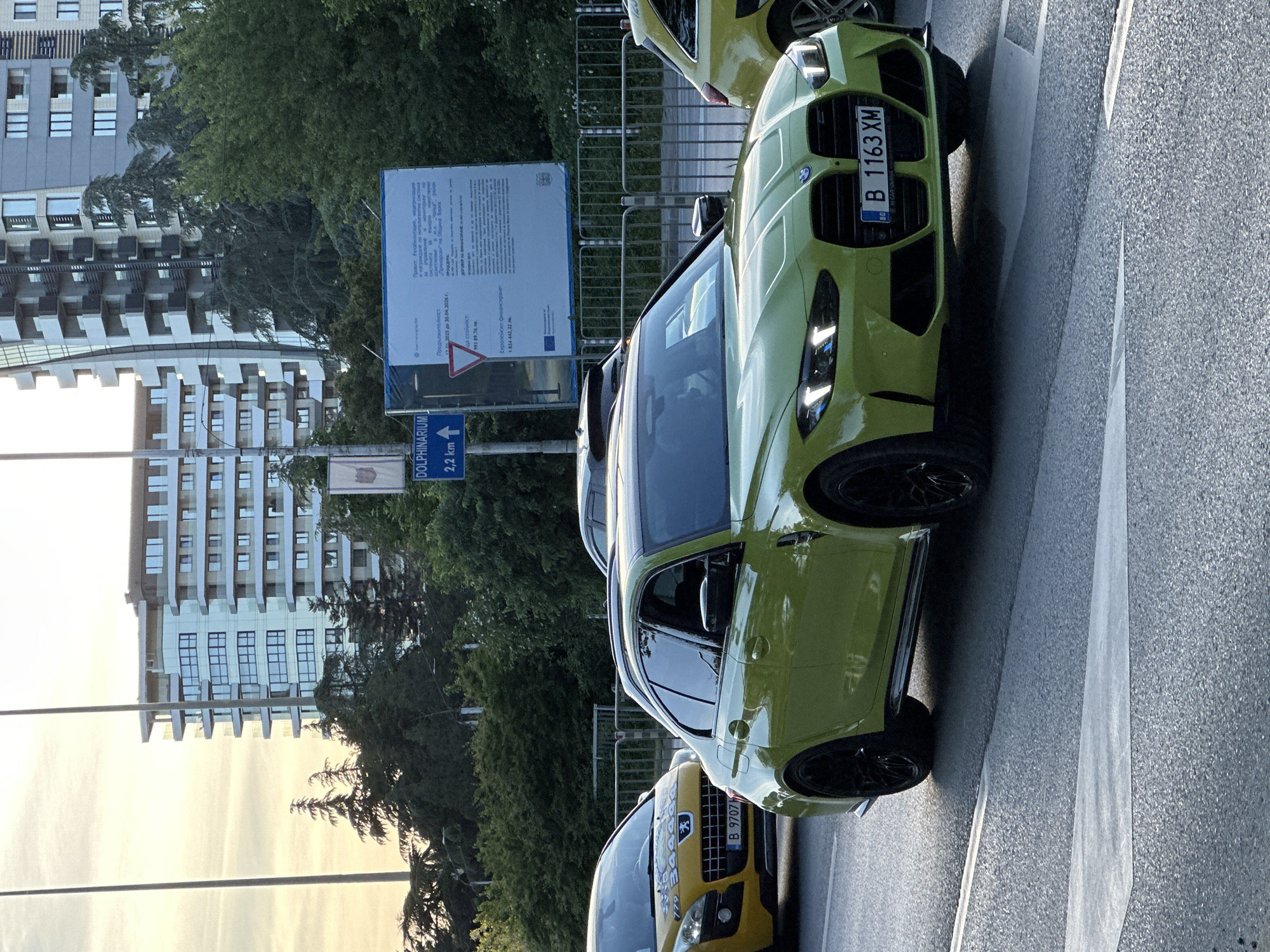 The image shows a street scene in Varna, Bulgaria. In the foreground, there are two cars parked on the side of the road. The car closest to the viewer is a lime green BMW with the license plate B 1163 XM. Behind it is a yellow Peugeot with the license plate B 9707. A sign indicates "Dolphinarium 2.2 km," suggesting the location is near the Varna Dolphinarium. Next to the road there is another sign indicating information about a municipal restoration project. In the background, there is a building and trees. The image appears to be taken during daylight hours.