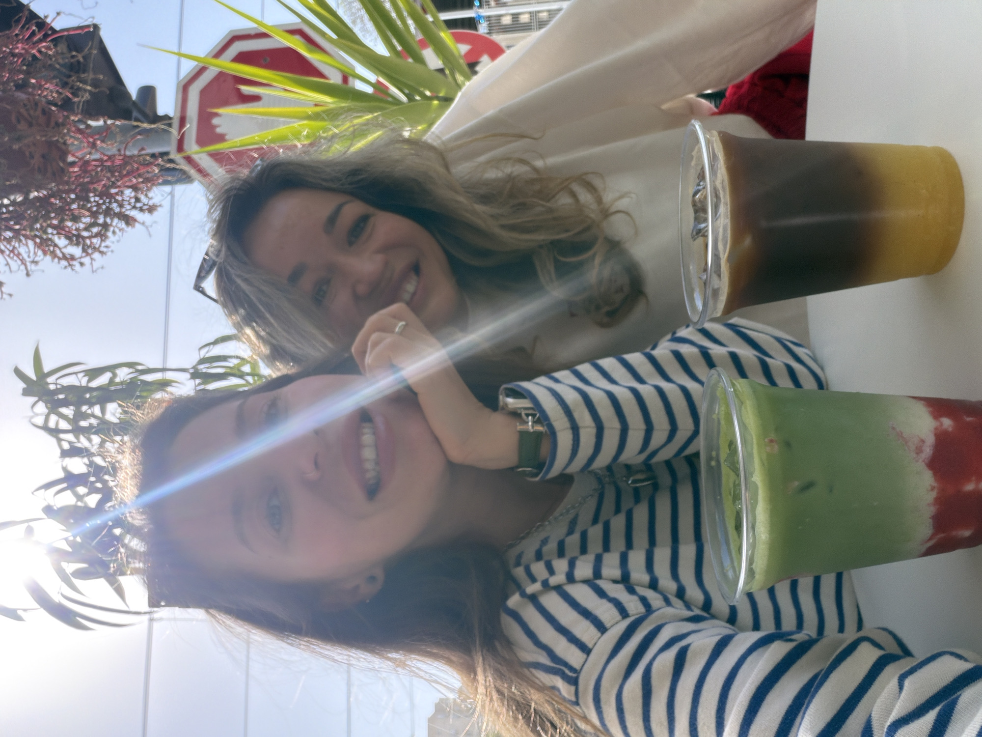 This is a bright, outdoor selfie featuring two young women enjoying drinks, likely at a cafe in Bat Yam, Israel. The image is taken from a low angle and is rotated on its side.
In the foreground, a woman with brown hair wears a white and blue striped long-sleeved shirt and a watch with a green strap on her left wrist. She is smiling broadly at the camera, with a sun flare partially obscuring her face. Behind her and slightly to the right, another woman with lighter, possibly blonde or light brown hair, also smiles at the camera. She is wearing a light-colored (possibly white or cream) top. Both women appear happy and relaxed.
On a white table in front of them are two colorful layered drinks in clear plastic cups. One drink features a dark brown layer (likely coffee) over a yellow or orange layer. The other drink has a vibrant green layer (possibly matcha) on top of a red layer (perhaps a fruit puree like strawberry).
The background shows lush green foliage and plants, suggesting an outdoor setting. Bright sunlight creates noticeable lens flares across the image, indicating a sunny daytime. Partially visible in the upper background, and appearing upside down due to the image rotation, is a red octagonal stop sign with white lettering. The word "STOP" is partially discernible on the sign.