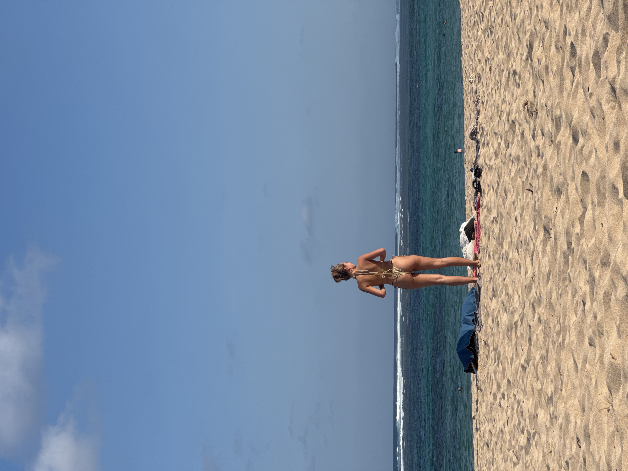 A woman stands on a sandy beach, facing away from the camera and towards the ocean in Kuta Selatan, Indonesia. She is wearing a gold bikini. The ocean has calm waves and is a deep blue-green color. There are a few scattered items on the sand, including a blue tarp, towels, and clothing. The sky is clear and blue with a few wispy clouds.