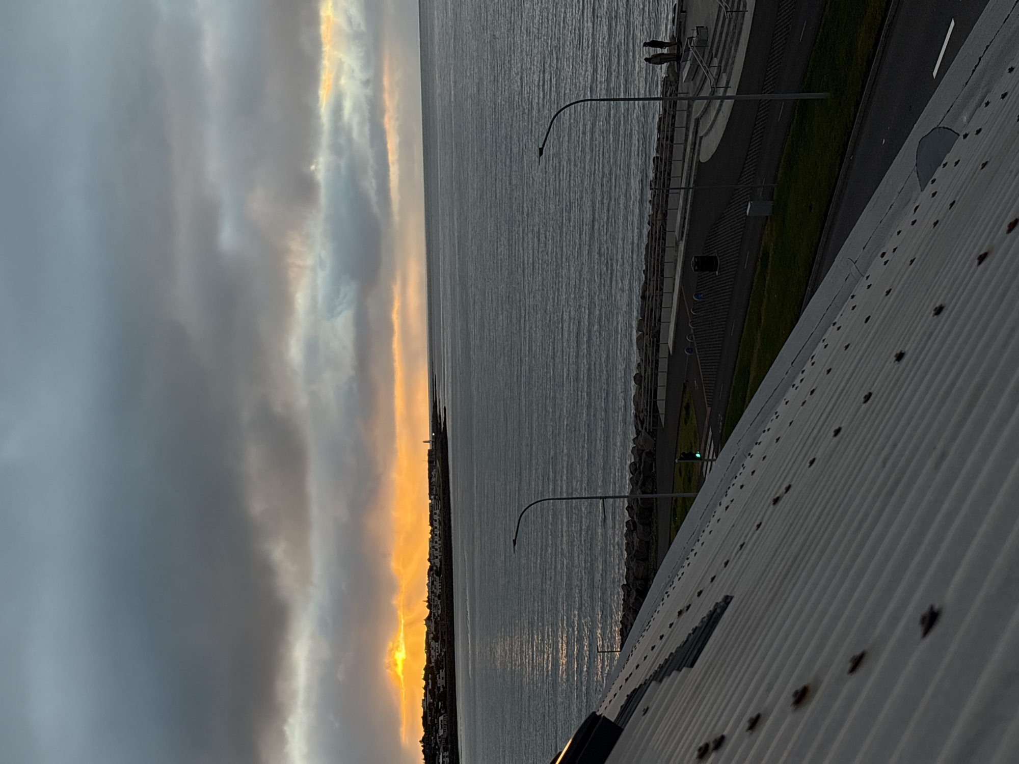 The image depicts a coastal view of Reykjavik, Iceland, at sunset. The foreground is dominated by the corrugated metal roof of a building, angled to the right. Beyond the roof, the scene opens to the ocean, with calm water reflecting the muted light.
Along the coastline, a paved walkway with bollards and seating areas is visible. Two figures stand on the walkway, facing the sea. Further along the coast, a line of low-rise buildings with dark roofs forms the Reykjavik skyline, silhouetted against the dramatic sunset. The sky is filled with dark, heavy clouds interspersed with streaks of bright orange and yellow light, indicating the sun's descent. A lighthouse is visible in the distance, positioned on the edge of the land. A traffic light showing green is visible on the road near the walkway. The overall atmosphere is one of a cool, possibly windy, evening.