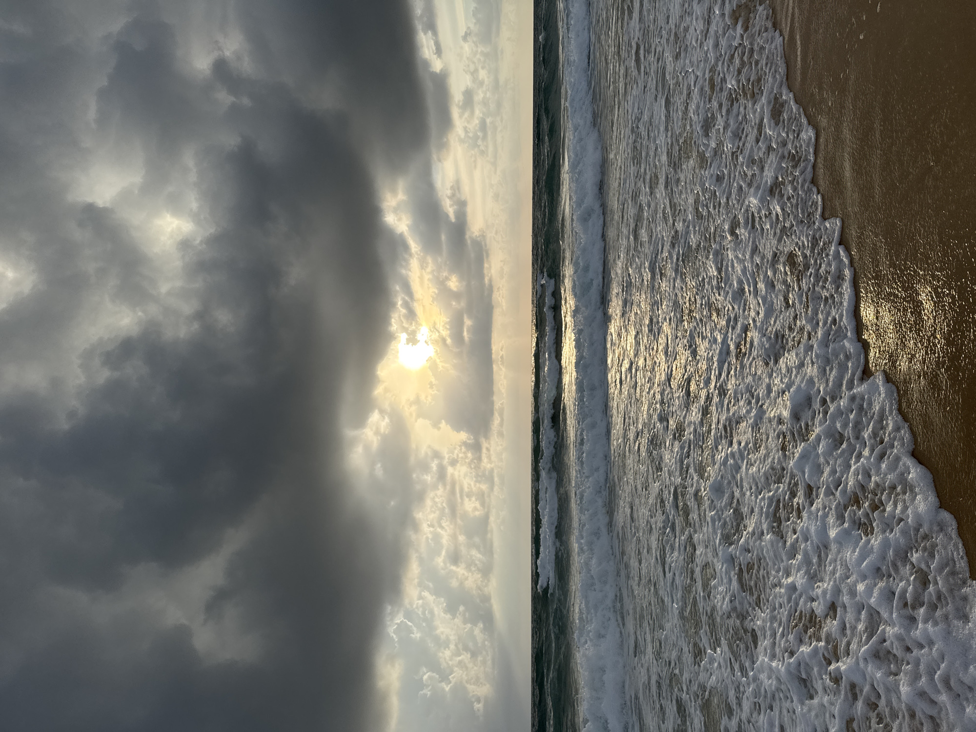 This is a vertical, eye-level, medium shot of a stormy beach scene in Karon, Thailand. The sky is dominated by dark, heavy clouds, but the sun is breaking through in the center, casting rays of light onto the water and sand. The ocean is a deep blue-green, with white foam from crashing waves that are reaching the sandy shore. The sand itself is a light brown color, wet from the receding tide and reflecting the sunlight. There are no people or objects visible in the frame.