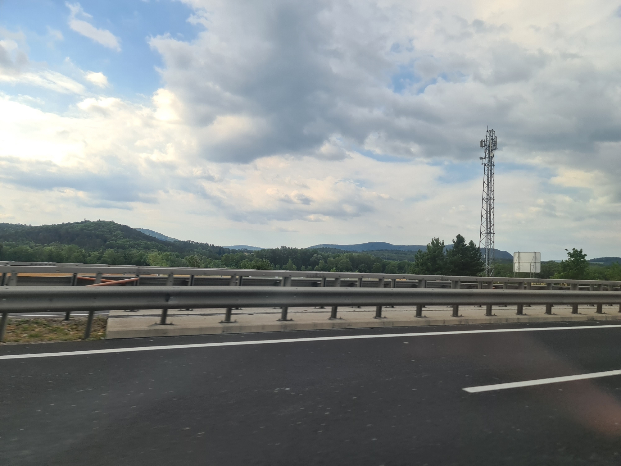 A wide shot shows a cell tower against a cloudy sky, with a lush green hillside and mountains in the background. In the foreground, a dark asphalt highway stretches into the distance, bordered by a metal guardrail and a white line. The scene appears to be on the outskirts of Breitenau, Austria.