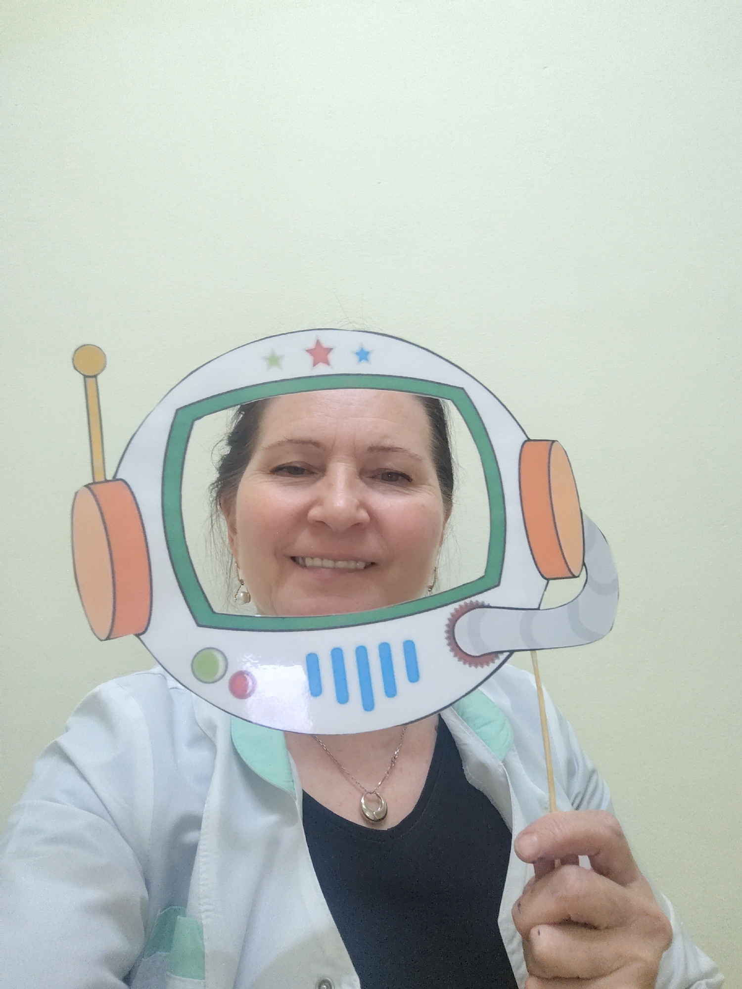 A woman is holding up a paper cut-out of an astronaut helmet, framing her face. She is wearing a white lab coat over a black shirt and a silver necklace with a round pendant. The helmet has an antenna on the left, a control panel with blue stripes at the bottom, and stars at the top. The background is a plain, light-colored wall. The location is Sarapul, Russia.