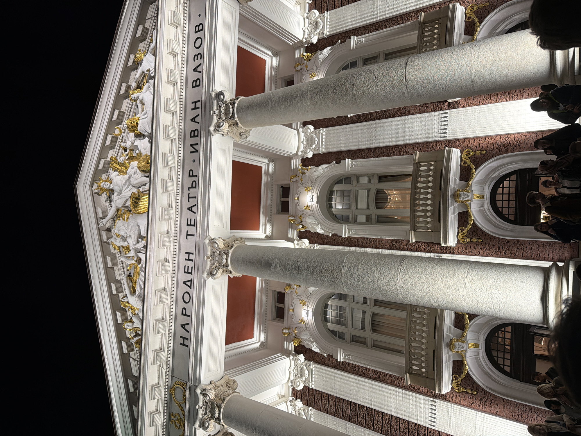 This image presents a low-angle, upward view of the illuminated facade of a grand building at night. The structure is identified as the Ivan Vazov National Theatre in Sofia, Bulgaria, by the prominent Cyrillic text "НАРОДЕН ТЕАТЪР · ИВАН ВАЗОВ" (National Theater · Ivan Vazov) vertically inscribed on a white frieze.
The building's architecture is classical, featuring imposing white columns with ornate Corinthian capitals. Above these, a triangular pediment is richly adorned with intricate white and gold sculptures depicting draped figures, possibly muses or mythological characters related to performance arts. The facade incorporates arched windows and balconies with delicate railings, set against a textured, dark red wall surface. Gold accents are visible on various decorative elements, including those beneath the balconies.
The scene is lit by artificial lights, casting a warm glow on the white and gold details and highlighting the building's architectural grandeur against the dark night sky. At the very bottom right, several indistinct human heads are visible, suggesting people are standing in front of the theater, looking up at its illuminated splendor. The photograph conveys a sense of the theater's majesty and cultural importance during the evening.