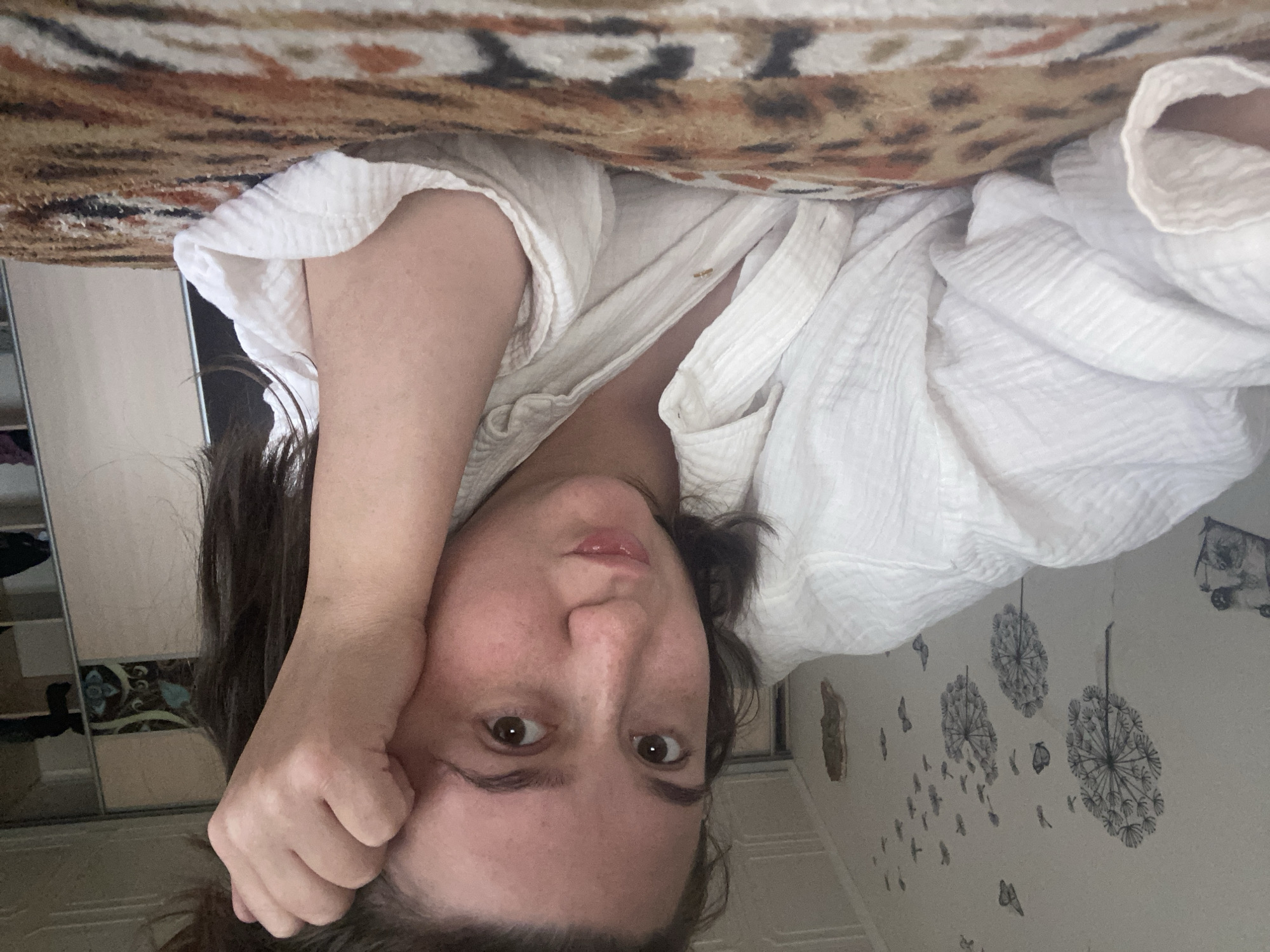 A young woman with dark hair and brown eyes is lying on a bed, propped up on her arm. She is wearing a white, button-down shirt that is slightly unbuttoned. The room appears to be a bedroom, with a closet visible in the background. The wall behind her is decorated with black dandelion and butterfly decals. The bedspread has a leopard print pattern. The overall setting suggests a casual, domestic environment, possibly a bedroom in Chaikovsky, Russia, given the provided context.