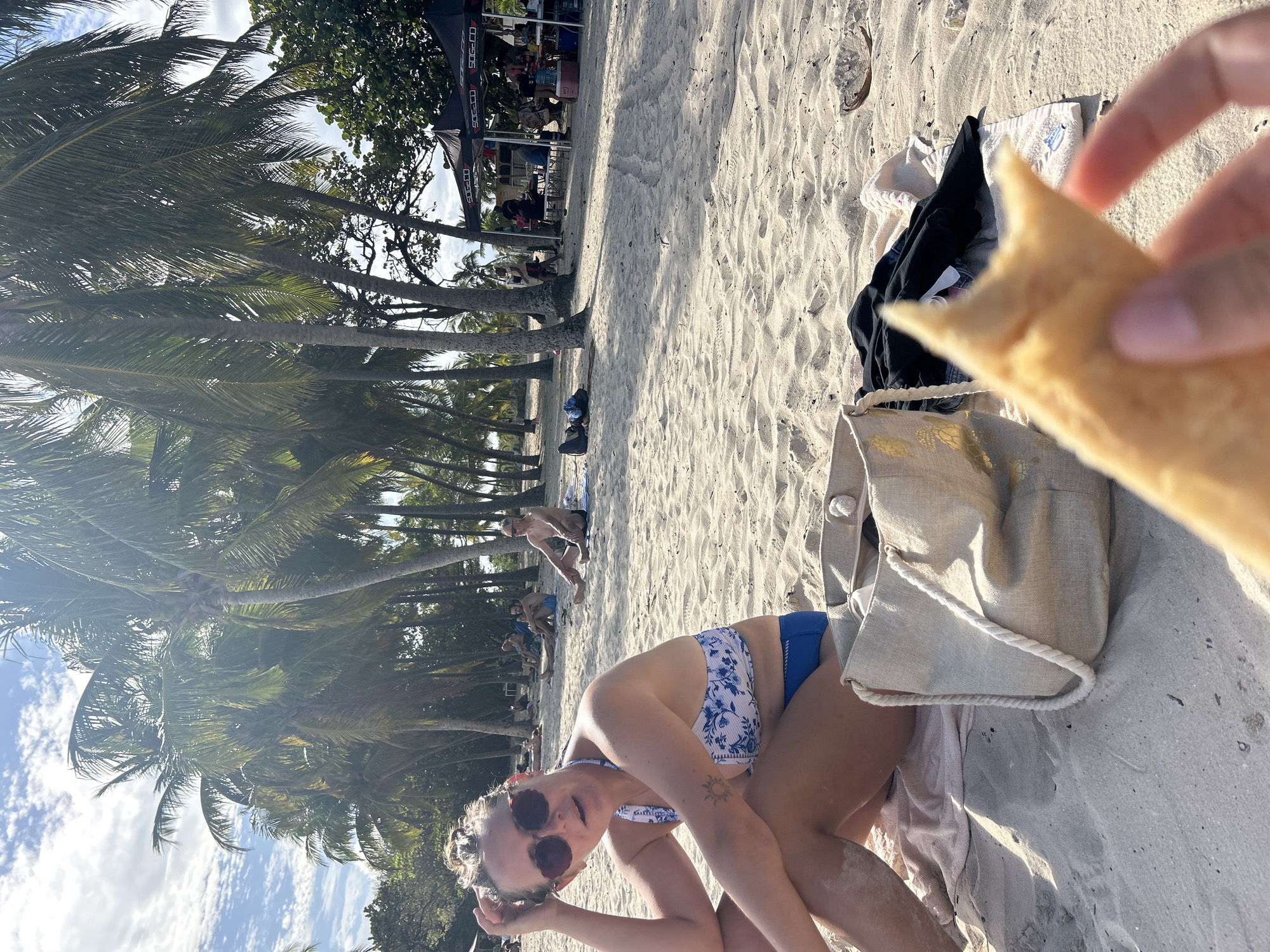 A medium shot captures a woman in a blue and white bikini, squatting on a sandy beach. She wears sunglasses and has a tattoo on her left arm. Her gaze is directed towards the viewer as she touches her hair. A hand holds a piece of food, partially obscuring the top right corner of the frame. Beside the woman sits a large tote bag on a beach towel, with other beachgoers visible in the background under the shade of palm trees. In the background, a building under a canopy with signage stands out. The beach is sandy, and the sky is visible through the palm trees. The scene is at Sainte-Anne, France.