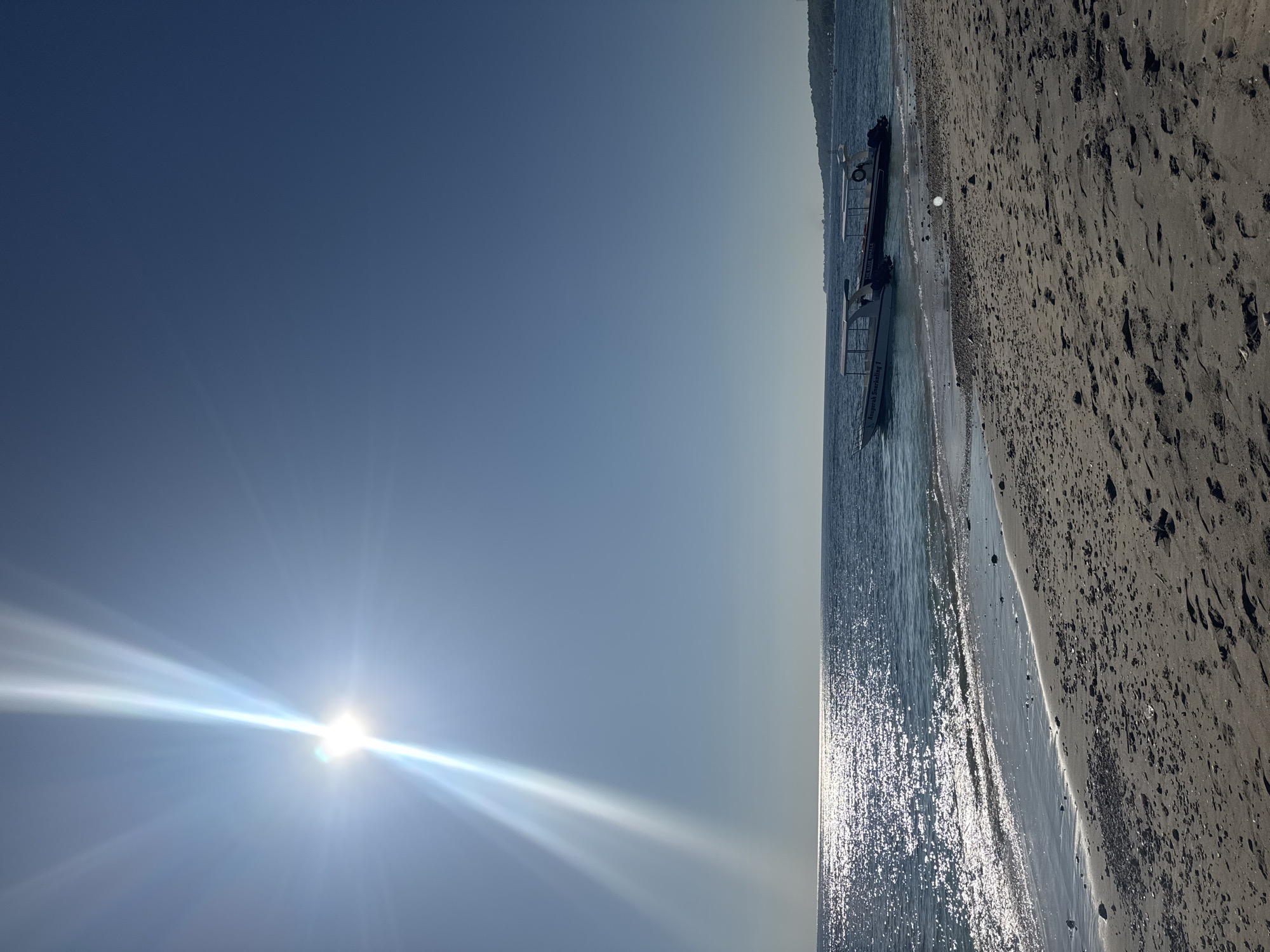 This image depicts a sunlit beach scene in Nusa Penida, Indonesia, under a clear blue sky. The intensely bright sun is positioned in the upper left quadrant, emitting strong starburst light rays and a prominent lens flare that streaks across the upper left and center of the frame. The ocean, visible across the middle ground, displays deep blue hues, with significant specular reflections from the sun shimmering on its surface, particularly towards the left. Gentle waves break onto a sandy beach in the foreground, where the light brown-grey sand is marked with numerous footprints and scattered small pebbles. Two small, white-canopied boats are partially beached or floating in shallow water on the right side of the image. The boat on the left is white and labeled "Anugerah Snorkeling 1," while the boat on the right is darker and inscribed "THE BLUE WHALE." A distant, vegetated landmass is visible on the horizon to the far right. No people are present in the scene.