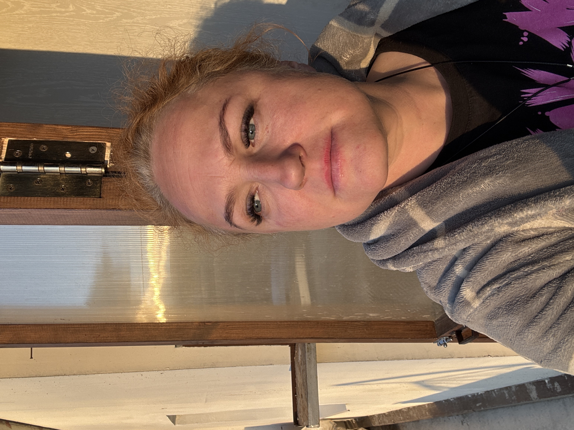 A woman is photographed indoors near a window. The location is presumed to be Chaikovsky, Russia. The woman has light skin, light brown hair, and blue eyes. She is wearing a dark-colored top with a purple design and a gray hooded sweatshirt. The window frame is brown and has a metal hinge visible in the upper left corner. The light outside the window creates a bright glow. The woman is facing the camera with a neutral expression. There are earphones visible around her neck.