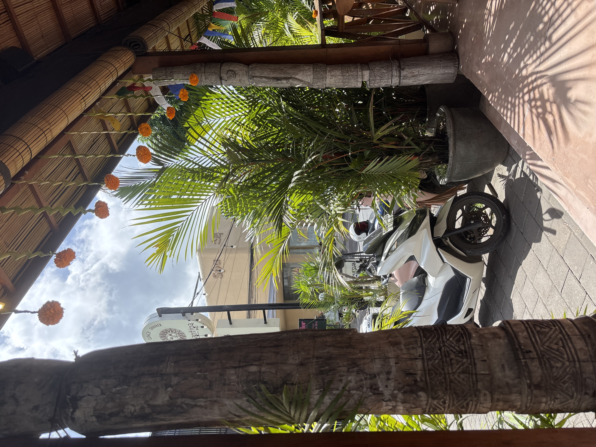 The photograph was taken in Kuta Selatan, Indonesia, from under an open-air structure with a bamboo and wood frame decorated with orange floral garlands and prayer flags. Through the frame, a scene unfolds of a sunlit street with lush green tropical plants and palm trees. The foreground is dominated by a thick, carved wooden beam. In the middle ground, a parked white motorbike is partially visible amongst the foliage. Beyond the greenery, buildings line the street, one with a sign that reads "Cantina." The sky is partly cloudy. The architecture and vegetation suggest a warm, tropical environment. Shadows cast by the plants dapple the ground and the wall of a building to the right.