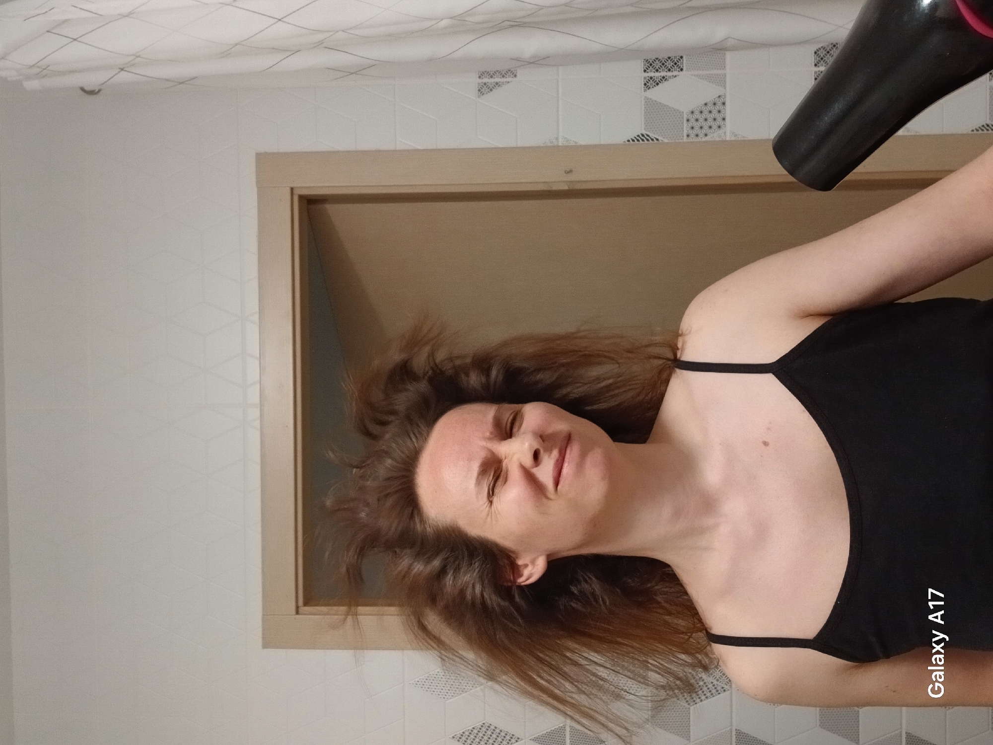 A woman with long brown hair is drying her hair with a black hairdryer. She is wearing a black tank top. She is in a bathroom with white walls and patterned tiles. A doorway is visible in the background. The location of the image in Yekaterinburg, Russia, cannot be determined.