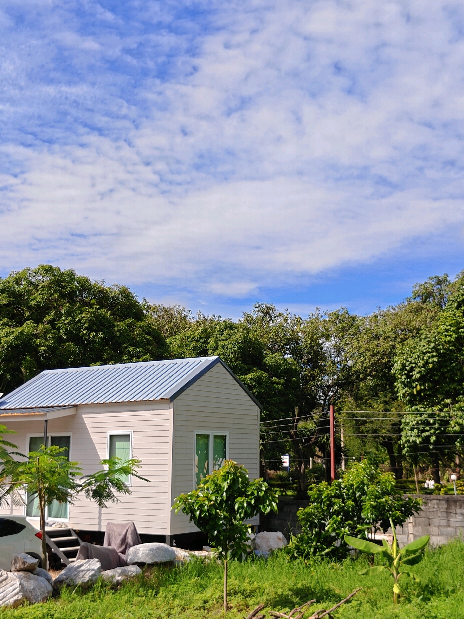 A small, modern, beige-colored house with a corrugated metal roof stands in a grassy area with trees and a blue sky with white clouds overhead. The house has multiple windows and a set of stairs leading to the front door. To the left of the house, a white car is parked. In front of the house, there are large rocks and dense green grass. To the right of the house, more trees and greenery are visible, along with a concrete wall and a small banana plant. The overall scene suggests a rural or suburban setting in Khao Yai, Thailand.
