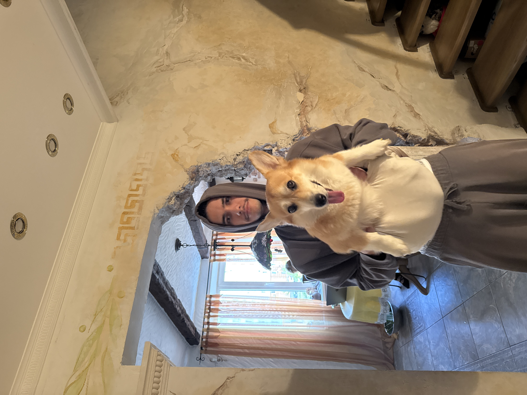 A man in a grey hooded sweatshirt is holding a corgi dog. The dog is sticking its tongue out. The scene is inside a building with ornate architectural details, including a decorative archway and exposed wooden beams. Sunlight streams in through a window in the background. There is no verifiable evidence that this photograph was taken in the city of Sevastopol, Ukraine.