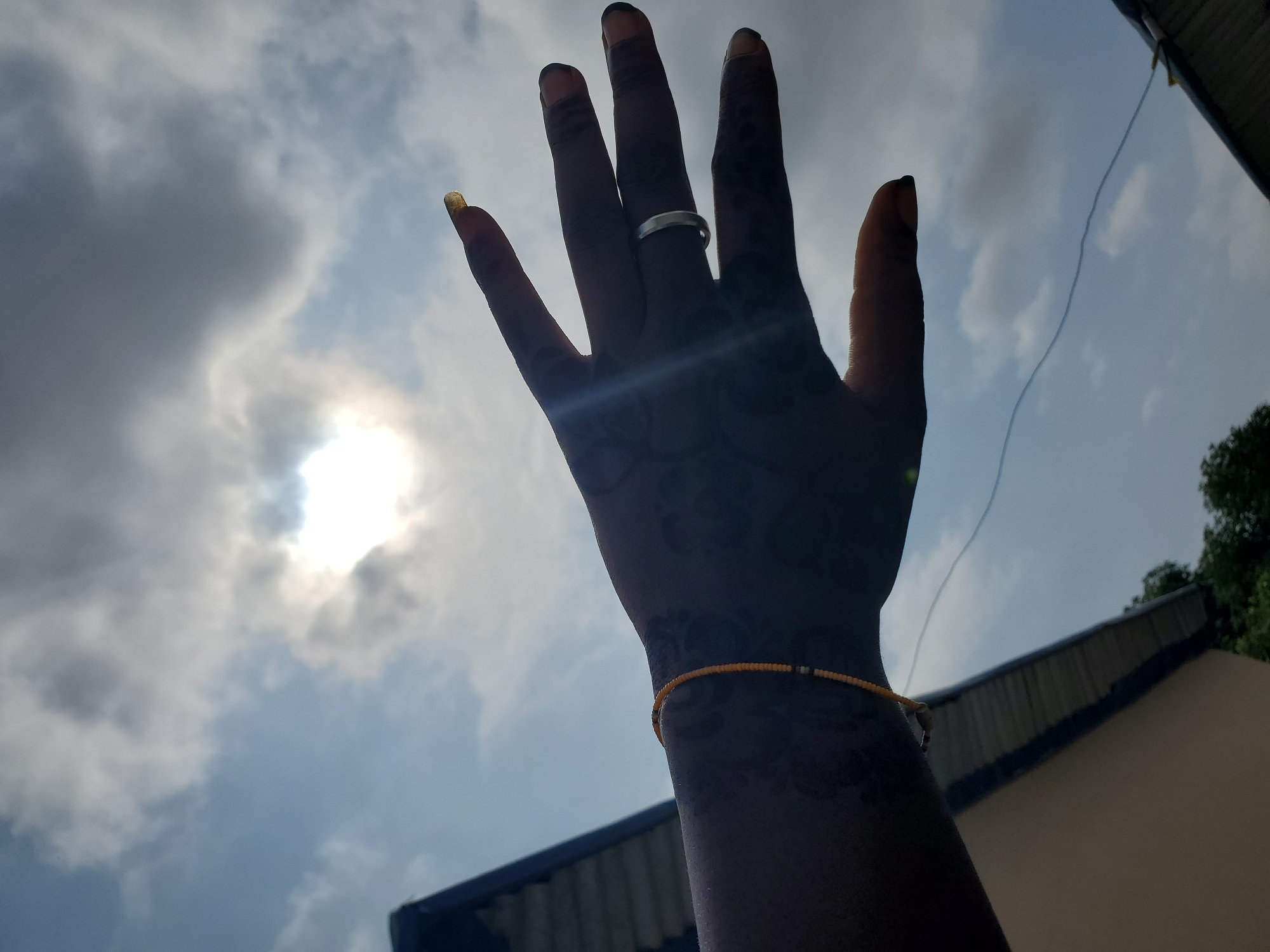 A close-up, low-angle shot captures a silhouetted dark-skinned hand raised against a bright, cloudy sky. The sun is partially obscured by clouds, creating a strong lens flare across the image. The hand has dark nail polish on some fingers, and a light-colored ring is visible on the middle finger. A delicate beaded bracelet, orange and black, adorns the wrist, and intricate henna patterns are visible on the back of the hand and forearm. In the background, the roof of a building with a corrugated metal design is visible, along with some green foliage and a thin wire strung across the scene. The lighting suggests it is daytime, possibly late afternoon due to the angle of the sun and the contrast between light and shadow. The overall impression is one of reaching or beckoning towards the sky. The location is Kason, Nigeria.