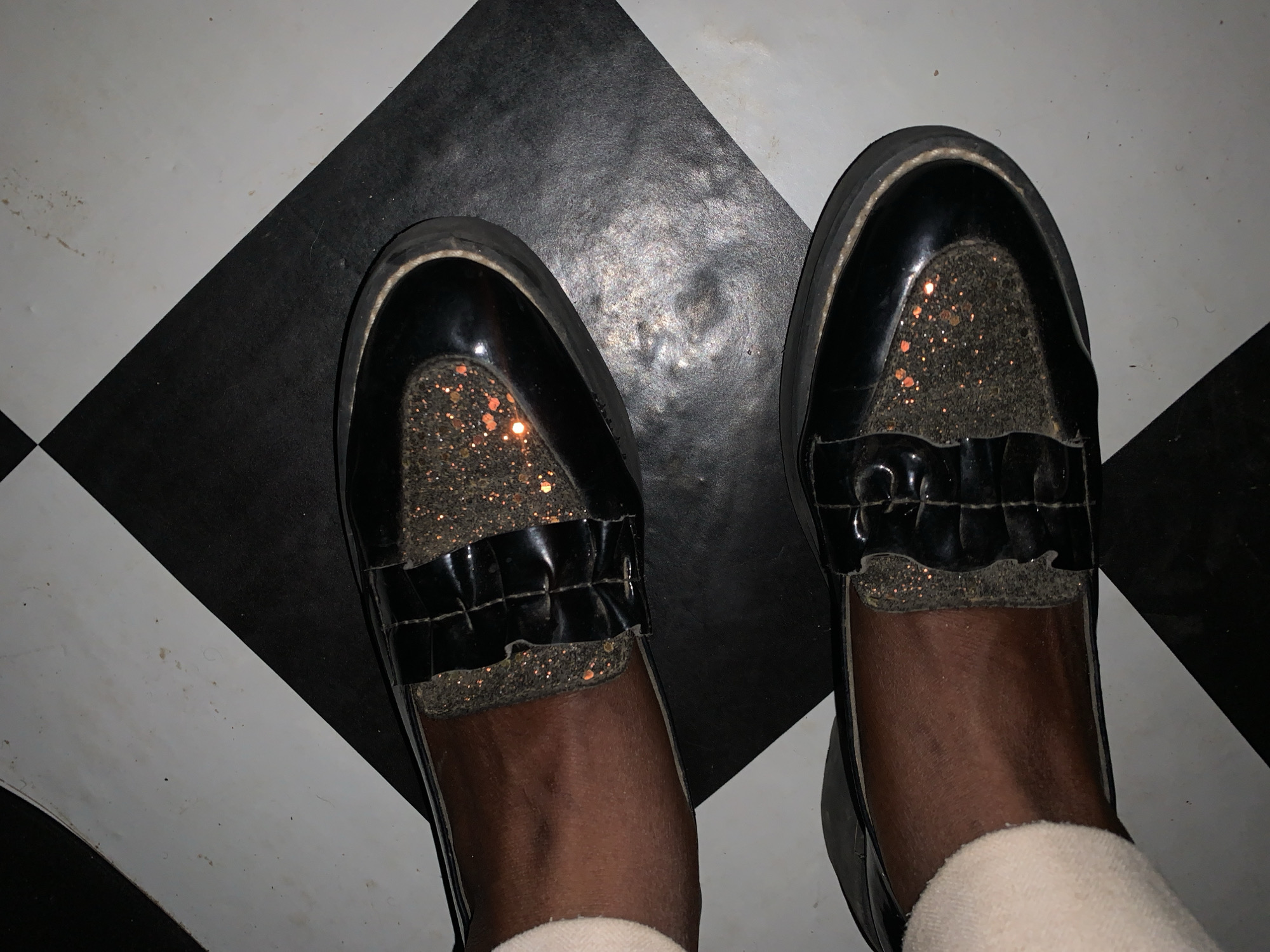This top-down view, taken in Rafin Par, Nigeria, captures a person's lower legs and feet wearing black platform shoes on a checkered floor. The subject has dark-toned skin, and white or cream-colored socks are visible around the ankles, peeking out from the shoes.
The footwear consists of black loafers or flat shoes with a glossy, patent leather-like finish. A distinctive feature is the vamp (upper front part) of the shoes, which is adorned with a dark gold or bronze glitter material, interspersed with brighter orange and gold speckles. A black, ruffled, or strapped detail, made of the same shiny material as the rest of the shoe, crosses this glitter section. The shoes have thick, black platform soles.
The setting is an indoor space with a black and white checkered floor, arranged diagonally beneath the feet. The white tiles show some faint scuff marks, and the black tiles also exhibit signs of wear, including lighter, scuffed areas that suggest reflections or accumulated dust. The lighting appears somewhat muted, emphasizing the glitter on the shoes and the sheen of their patent material. The activity depicted is a person standing or posing, showcasing their footwear. No text is visible in the image.