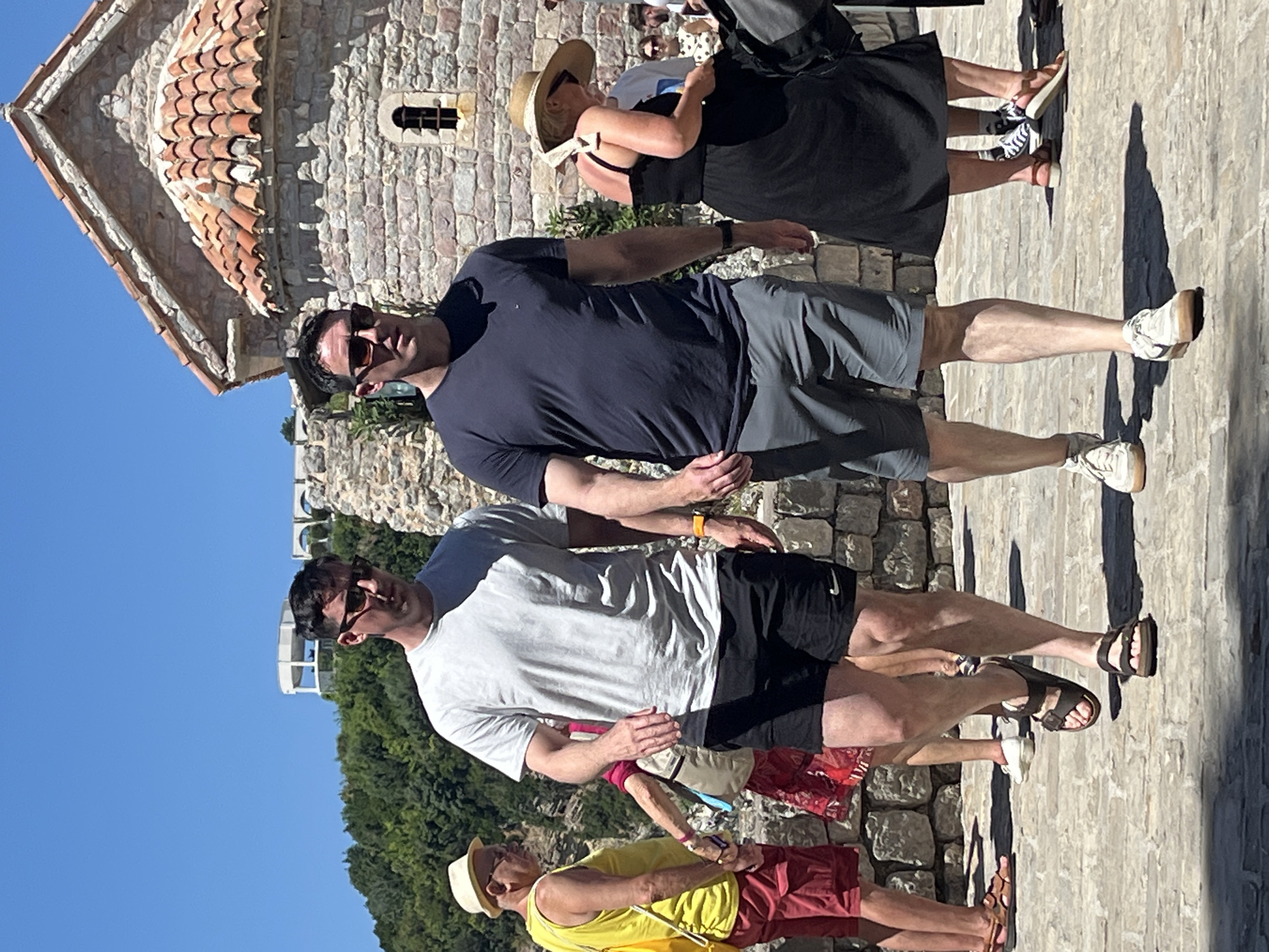 Two men are walking on a cobblestone path in front of a stone building with a red tiled roof. The man on the left is wearing a light grey t-shirt and black shorts, and the man on the right is wearing a dark blue t-shirt and grey shorts. Both men are wearing sunglasses. Behind them, a woman in a black dress and a straw hat is walking away from the camera. In the background, there is a lush green hillside with a white structure on top. The image appears to be taken in a historic part of Budva, Montenegro, possibly near the Church of St. Sava or on the grounds of the Budva Citadel. The lighting suggests it is a sunny day.