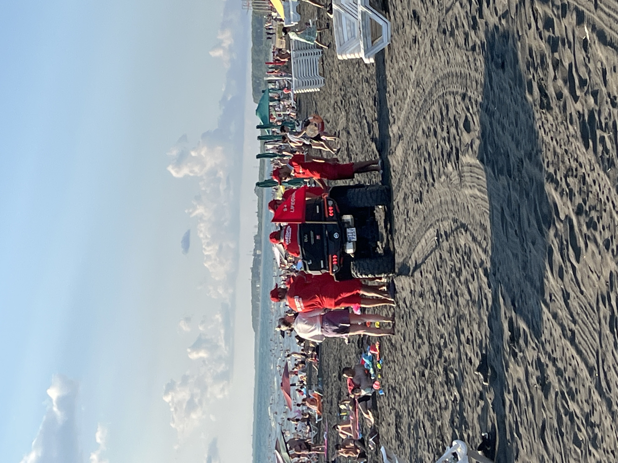 The image captures a scene on the black sand beach of Ureki, Georgia. In the foreground, an all-terrain vehicle (ATV) is parked on the sand, facing away from the camera. Three individuals in red lifeguard uniforms are standing near the ATV, with two of them looking back towards the water. One lifeguard is holding a red flag with "LIFEGUARD" printed on it. Behind the ATV, the beach is populated with numerous sunbathers and beachgoers, many of whom are seated on lounge chairs or towels under umbrellas. The Black Sea is visible in the background, with waves and a hazy coastline. The sky is partly cloudy with a bright, sunny atmosphere. The ground is composed of dark, fine-grained sand with tire tracks from the ATV imprinted on it. Several beach chairs and umbrellas are scattered across the beach, indicating a busy day at the resort.