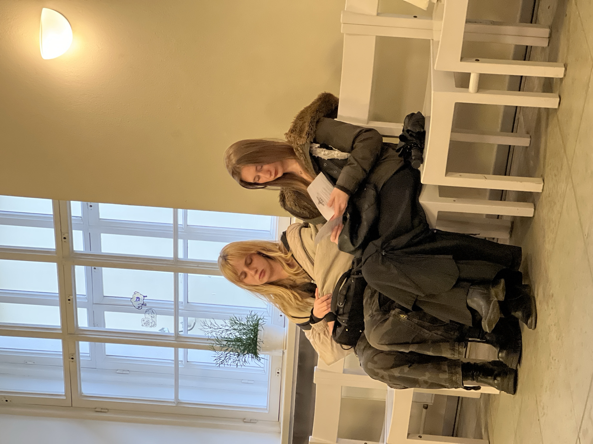 Two young women are sitting side by side on a white pew. One woman has brown hair and is wearing a dark coat with a fur-lined hood, holding a small booklet. The other woman has blonde hair and is wearing a beige coat. Both women appear to be looking down. They are indoors, with a large window to their left, revealing an outdoor scene. A small potted plant is visible on the windowsill. The location is likely a church or similar place of worship in Saint Petersburg, Russia. A white wall sconce is visible. There are cartoon stickers on the window.