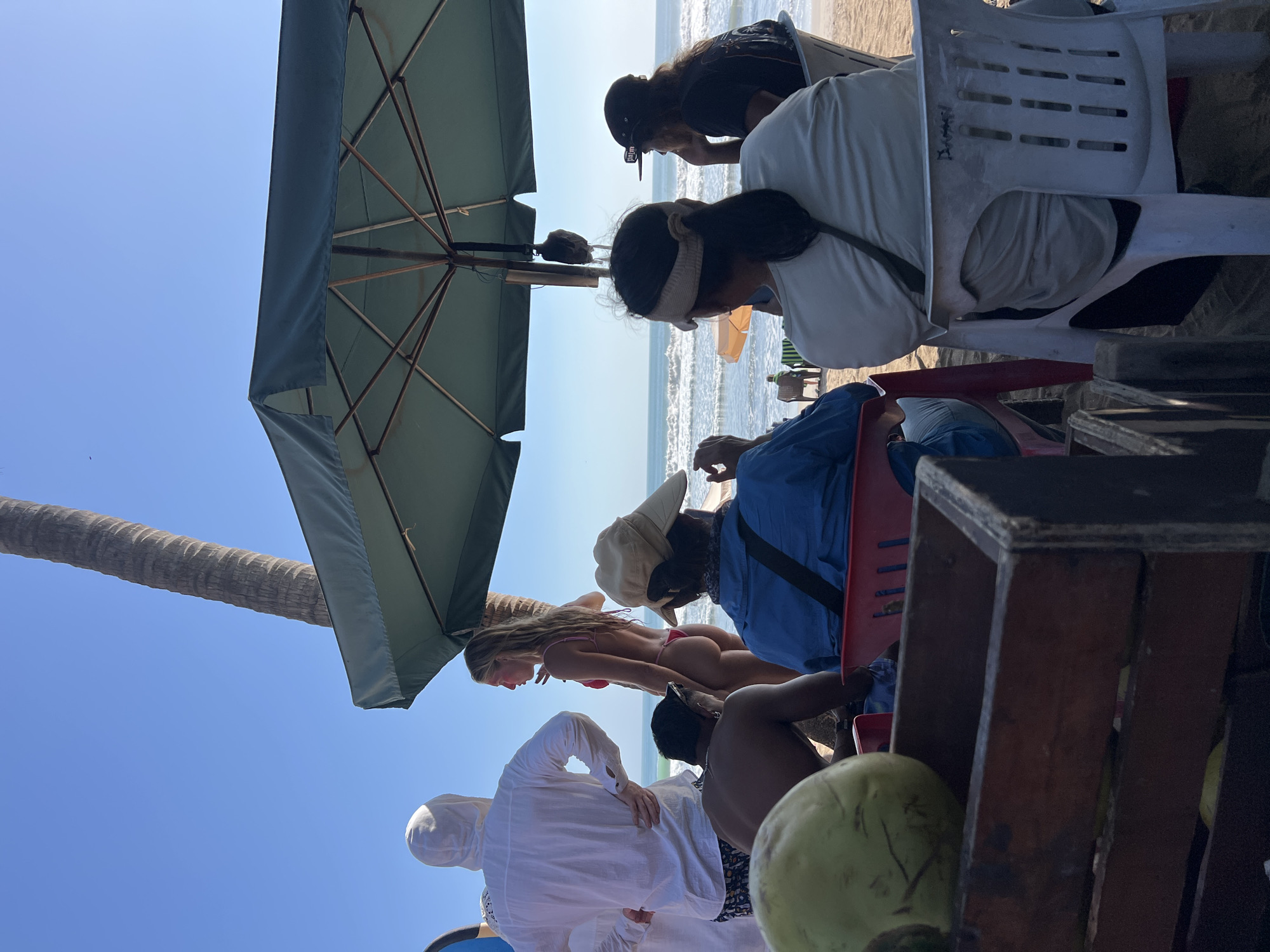 A group of people are gathered on a beach in Kuta, Indonesia, under a large green umbrella. The ocean with gentle waves is visible in the background. A tall palm tree stands to the left, its trunk visible against a clear blue sky. In the foreground, a wooden crate holds a green coconut.
Several individuals are seated or standing. A man in a white hooded jacket is looking towards the ocean, his back to the camera. A woman in a red bikini stands in profile, facing the water. Another man is seated in the foreground, facing away from the camera, wearing a blue shirt and a light-colored hat. To the right, two more people are seated under the umbrella, one wearing a white shirt and a visor, the other a black t-shirt and a baseball cap.
The atmosphere appears relaxed, suggesting a casual beach outing or a stop at a beachside vendor. The lighting indicates it is daytime, likely morning or afternoon, with the sun casting shadows on the sand.