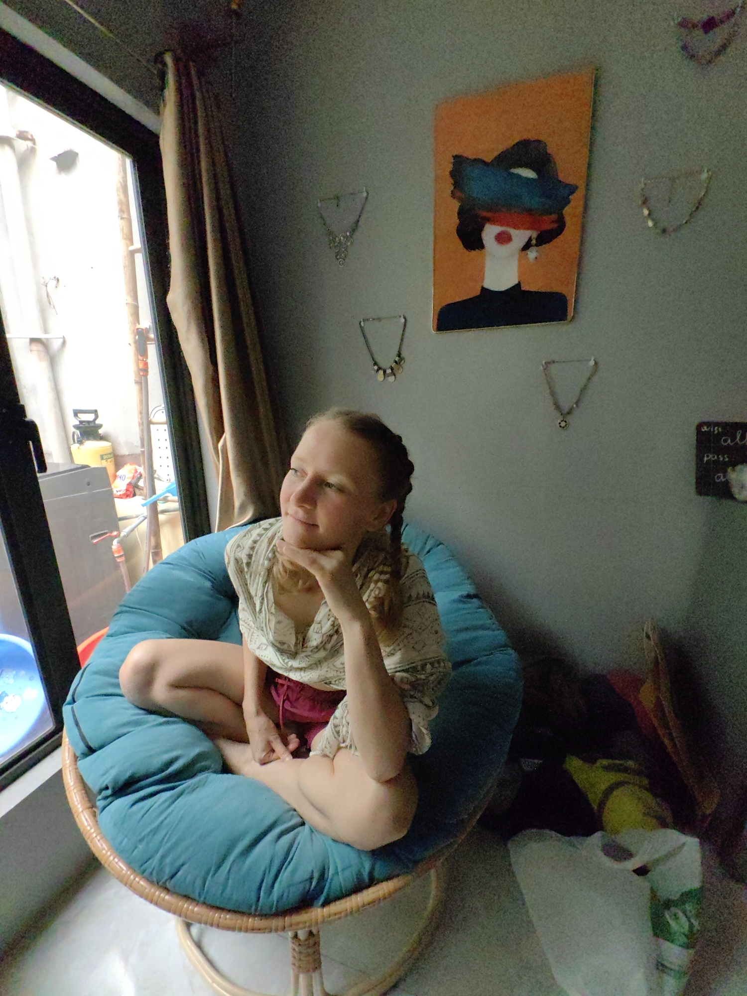 A medium shot shows a young woman seated in a blue wicker chair with a puffy cushion, indoors near a window. She is fair-skinned with blonde hair braided and wearing a light-colored patterned shirt and reddish shorts. She is looking off to the side with her hand resting on her chin, appearing thoughtful.
Outside the window, obscured by partial reflections, are hints of a residential or commercial area with stacked items and a bright yellow object resembling equipment. The room has gray walls decorated with jewelry and an orange portrait of a woman with a hat. In the corner, there is a cluttered area with bags and objects, suggesting a lived-in or storage space.
This scene is likely located in Hà Nội, Vietnam.