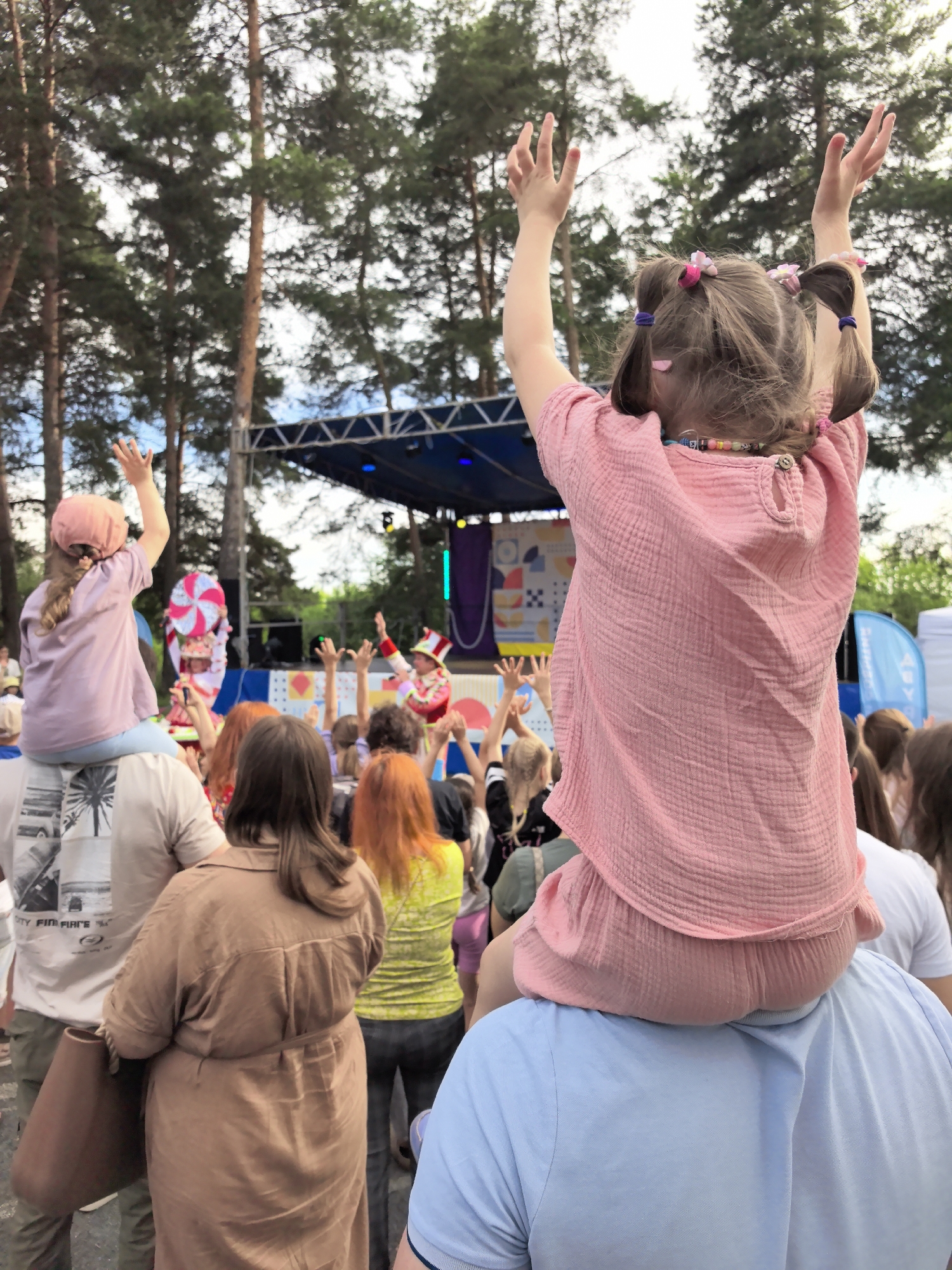 In the city of Gorniy, Russia, a crowd of people gather outdoors to watch a performance on a stage. The perspective is from behind the crowd, looking towards the stage. In the foreground, a young girl is being carried on someone's shoulders, her arms raised in excitement. She is wearing a pink outfit and her hair is styled in pigtails with colorful hair ties. To the left, another child is also being carried on shoulders, wearing a pink cap and a light purple shirt, with their arm raised. The stage is visible in the background with performers in colorful costumes, including one in a red and white jester-like outfit. The backdrop of the stage features a colorful abstract design. Trees with green foliage surround the area, indicating an outdoor setting, likely a park or festival grounds. The overall atmosphere appears to be festive and engaging.
