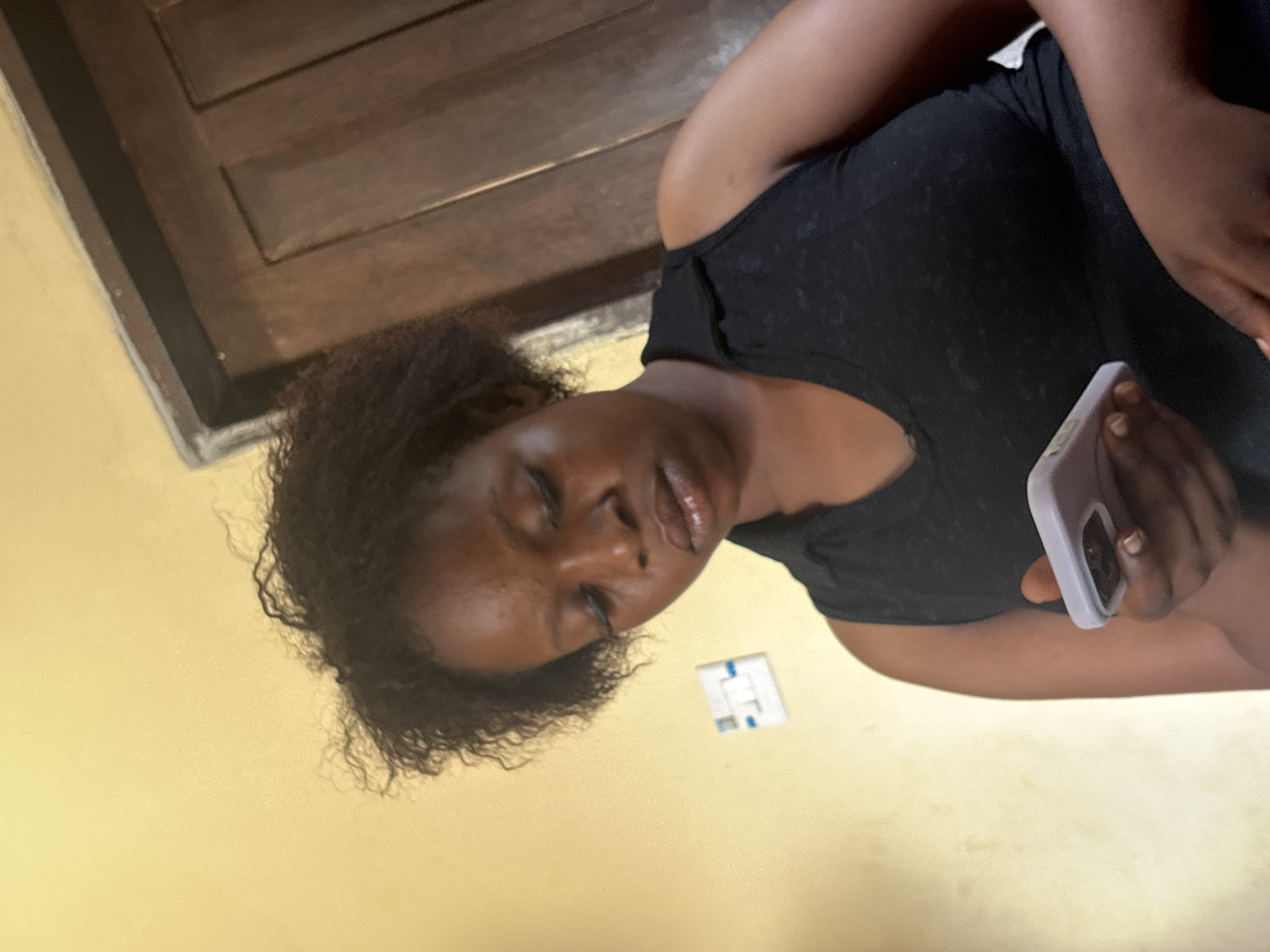 A close-up, tilted image captures a person with dark skin and curly dark hair, appearing to be a woman, in an indoor setting. She is seen from the chest up, wearing a dark (likely black) top. Her eyes are closed or heavily lidded, and her lips are slightly parted and appear glossy, conveying a relaxed or restful state.
The person holds a light-colored, possibly light purple or white, smartphone in her right hand, visible in the lower right of the frame. The background features a warm, light yellow wall, with a white electrical switch or outlet containing blue markings located to the left of her head. In the upper left background, there is a dark brown wooden structure, possibly part of a door or window frame, with horizontal paneling. The lighting suggests an interior space. No discernible text is visible in the image. The scene is consistent with an indoor environment in Ikot Ekpene, Nigeria.