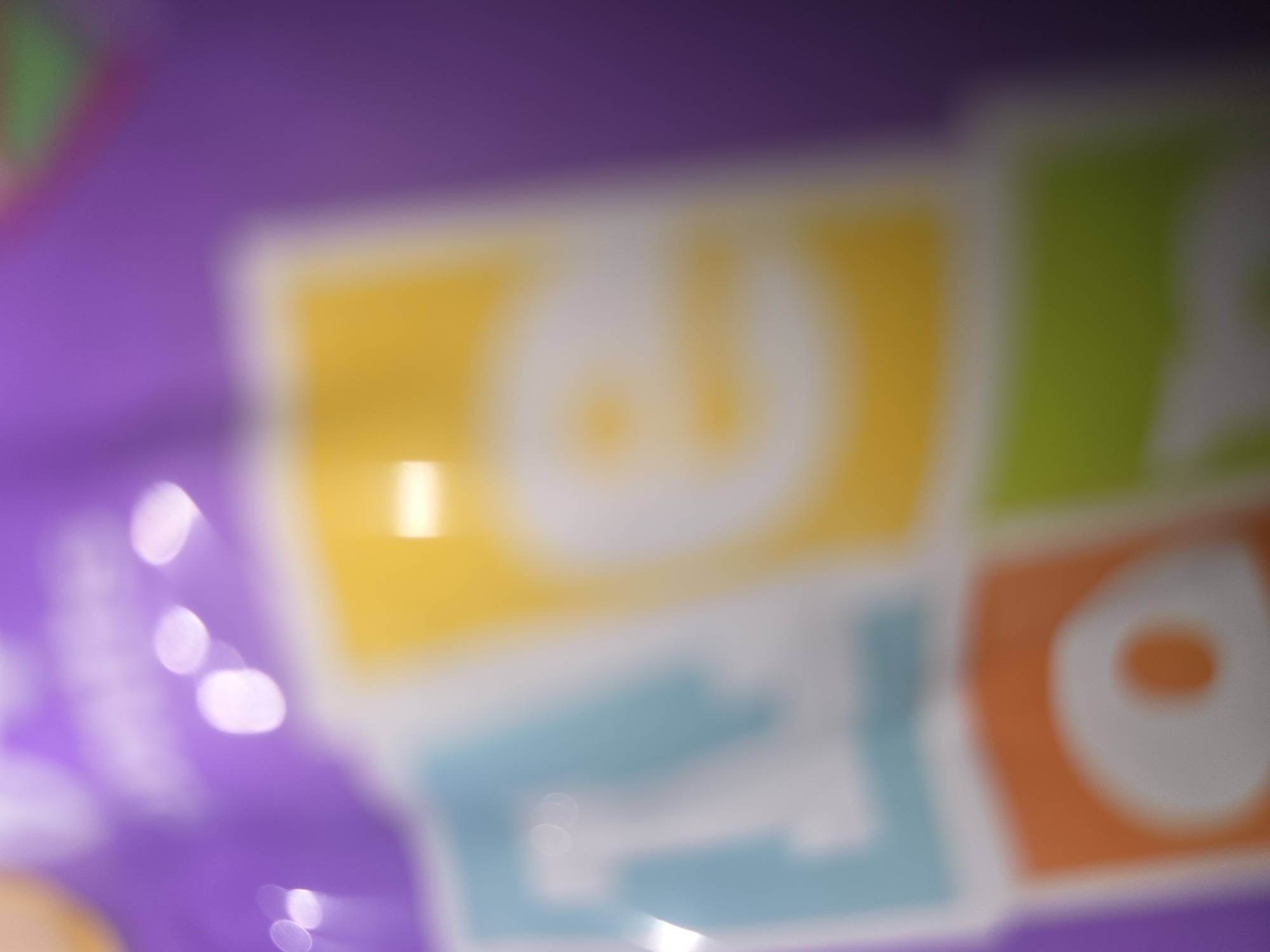 The image is a close-up, out-of-focus shot of colorful blocks with letters on a purple background. The blocks appear to be light blue, yellow, orange, and green. The letters on the blocks are white and blurred, but suggest a word or words. There are also several bright white circular highlights, likely reflections on the surface of the blocks or packaging. The overall impression is of a children's toy or game. The image does not contain any recognizable landmarks or contextual clues that would allow for identification of its location in Ulan-Ude, Russia, or any other specific place.