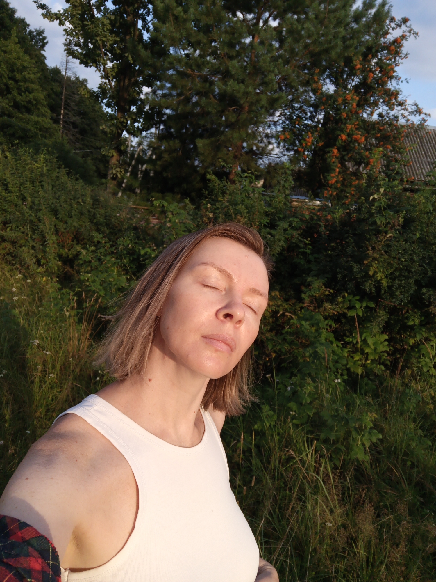 An adult female with light brown hair is captured in a close-up, selfie-style shot outdoors. She is wearing a white tank top, and her eyes are closed, with her head tilted slightly upwards as if enjoying the sunlight. Her skin appears flushed, likely from the sun. The background consists of lush green foliage, tall grass, and trees, with a hint of a grey roof visible through the leaves in the upper right corner, suggesting a rural or semi-rural setting. The lighting indicates it is either early morning or late afternoon. The scene does not contain any visible elements that specifically identify the location as Chaikovsky, Russia.