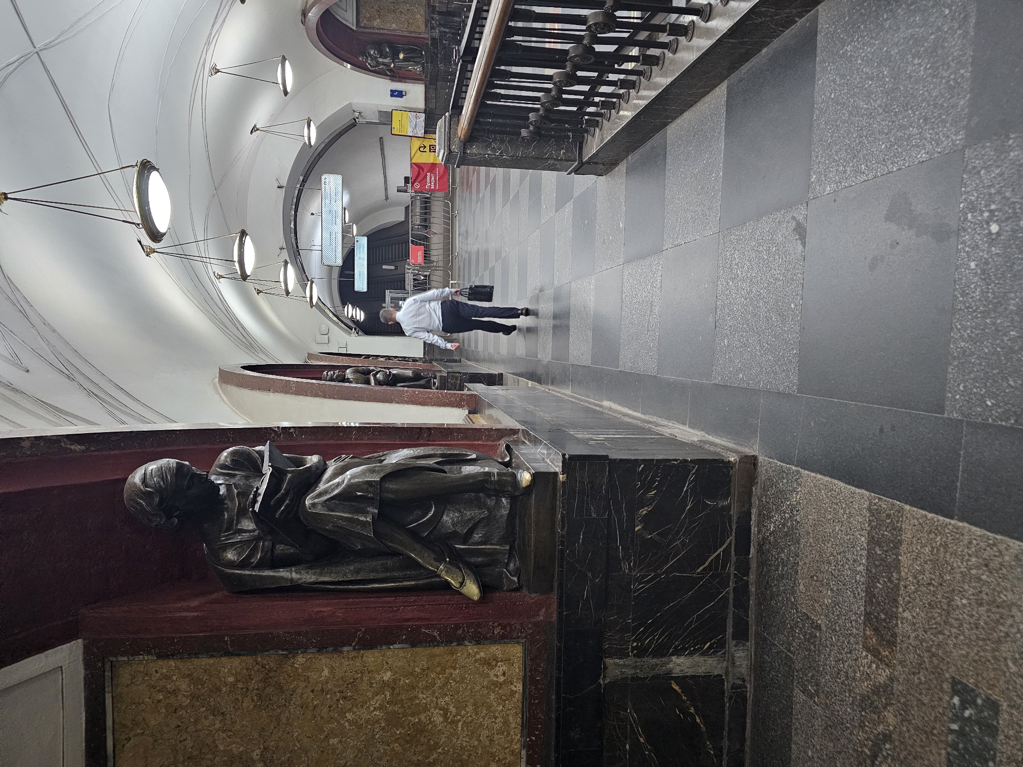 The image captures a view inside a Moscow Metro station, specifically the Komsomolskaya (Koltsevaya Line) station, known for its ornate architecture. On the left, a bronze sculpture of a woman reading is prominently displayed within a niche. The sculpture is dark and detailed, with the woman depicted in a seated pose, engrossed in a book. Adjacent to the sculpture is a wall with a textured, marble-like finish. The floor is paved with large, dark grey and black square tiles, some with a speckled granite texture.
In the mid-ground, a man in a light-colored dress shirt and dark trousers walks away from the camera, carrying a black briefcase in his right hand. He is proceeding down the tiled walkway. The environment of the station features an arched ceiling with numerous pendant lights, characteristic of the station's grand design. Various signs and barriers are visible further down the corridor, indicating different areas or temporary closures. The overall atmosphere is one of subdued activity within a richly decorated public space.
