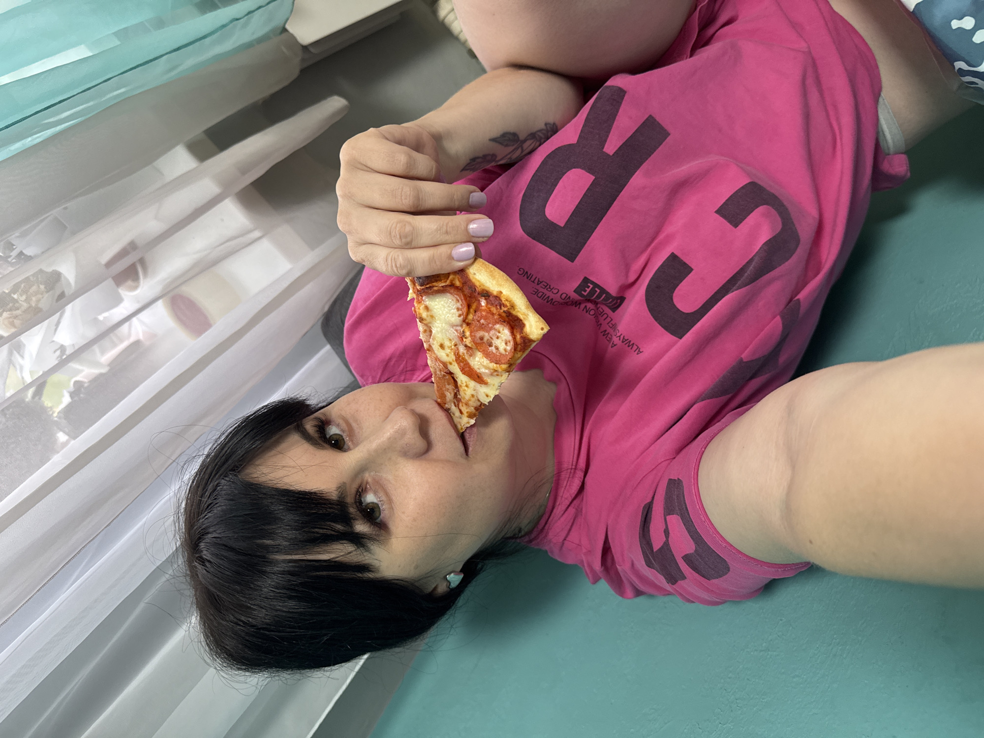 A woman with dark hair and bangs is lying down and eating a slice of pizza. She is wearing a pink t-shirt with dark lettering. Her nails are painted a light purple color. The background includes a window with white curtains, a white wall, and a light green surface. The image was taken in the city of Zav'yalovo, Russia.