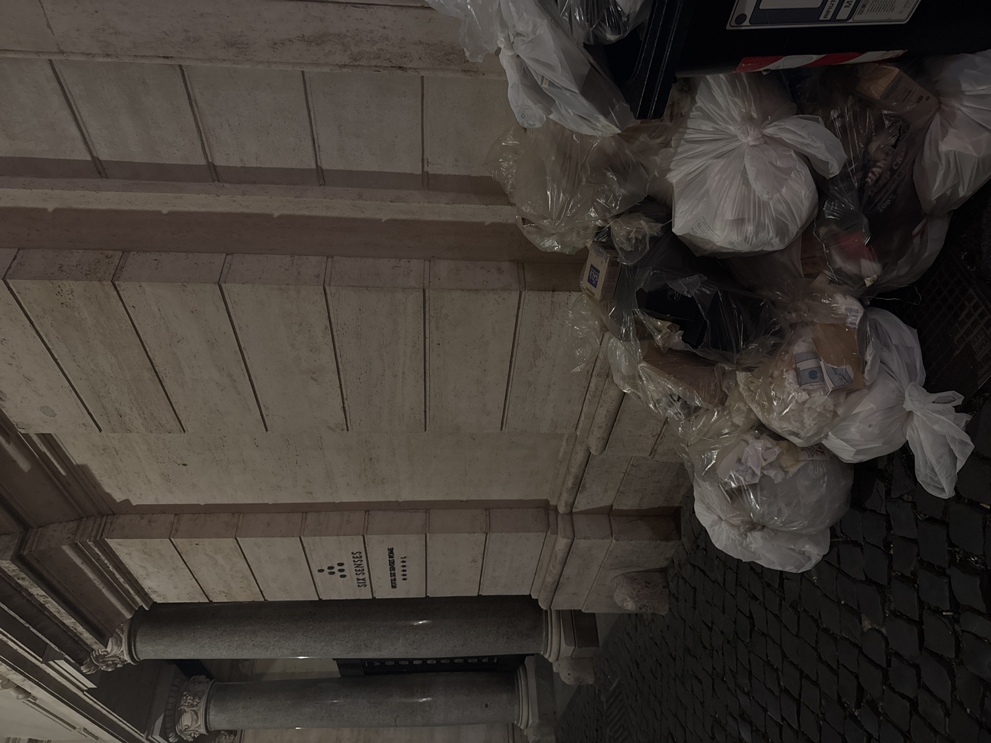 The image captures a scene in Rome, Italy, specifically at a location bearing the sign "SIX SENSES HOTEL SIX SENSES ROME." It shows a portion of an architectural structure, possibly an entrance or facade, supported by stone columns. The environment is urban, evidenced by the cobblestone ground and the presence of accumulated refuse bags and a trash bin overflowing with trash. The refuse consists of white and clear plastic bags, cartons, and other discarded items. The lighting suggests either night time or a dimly lit interior space. The image does not depict people.
