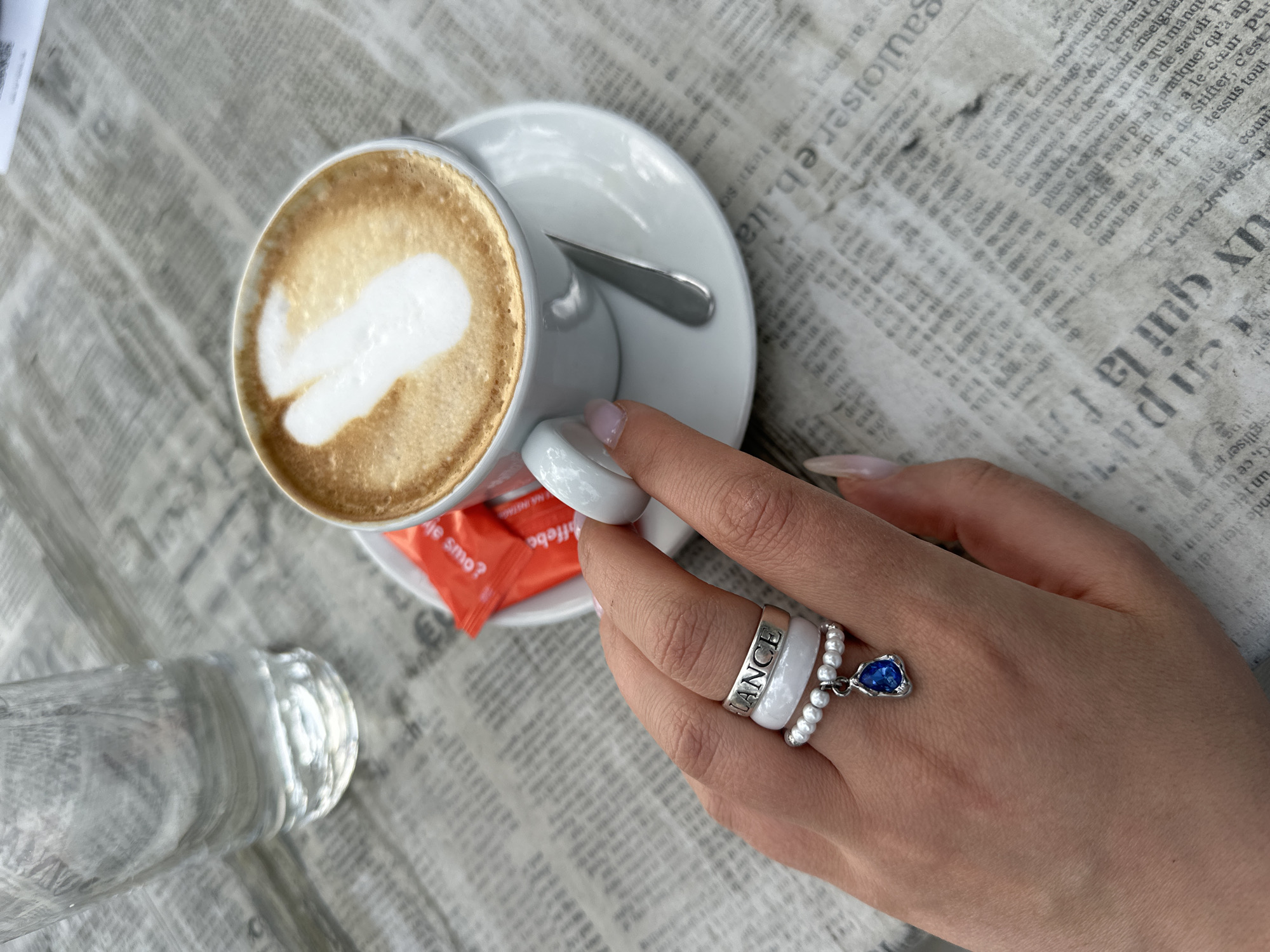 The image shows a top-down view of a cappuccino with latte art, on a white saucer with a spoon, being held by a person's hand. The person is wearing multiple rings: one with the word "LANCE", a ring made of white beads, and a ring with a blue teardrop gem. An orange sugar packet is visible beneath the saucer. A water bottle is partially visible in the lower left corner. The table is covered with a newspaper print. This scene likely takes place in Šušanj, Montenegro based on the text on the sugar packet, which is in Montenegrin.