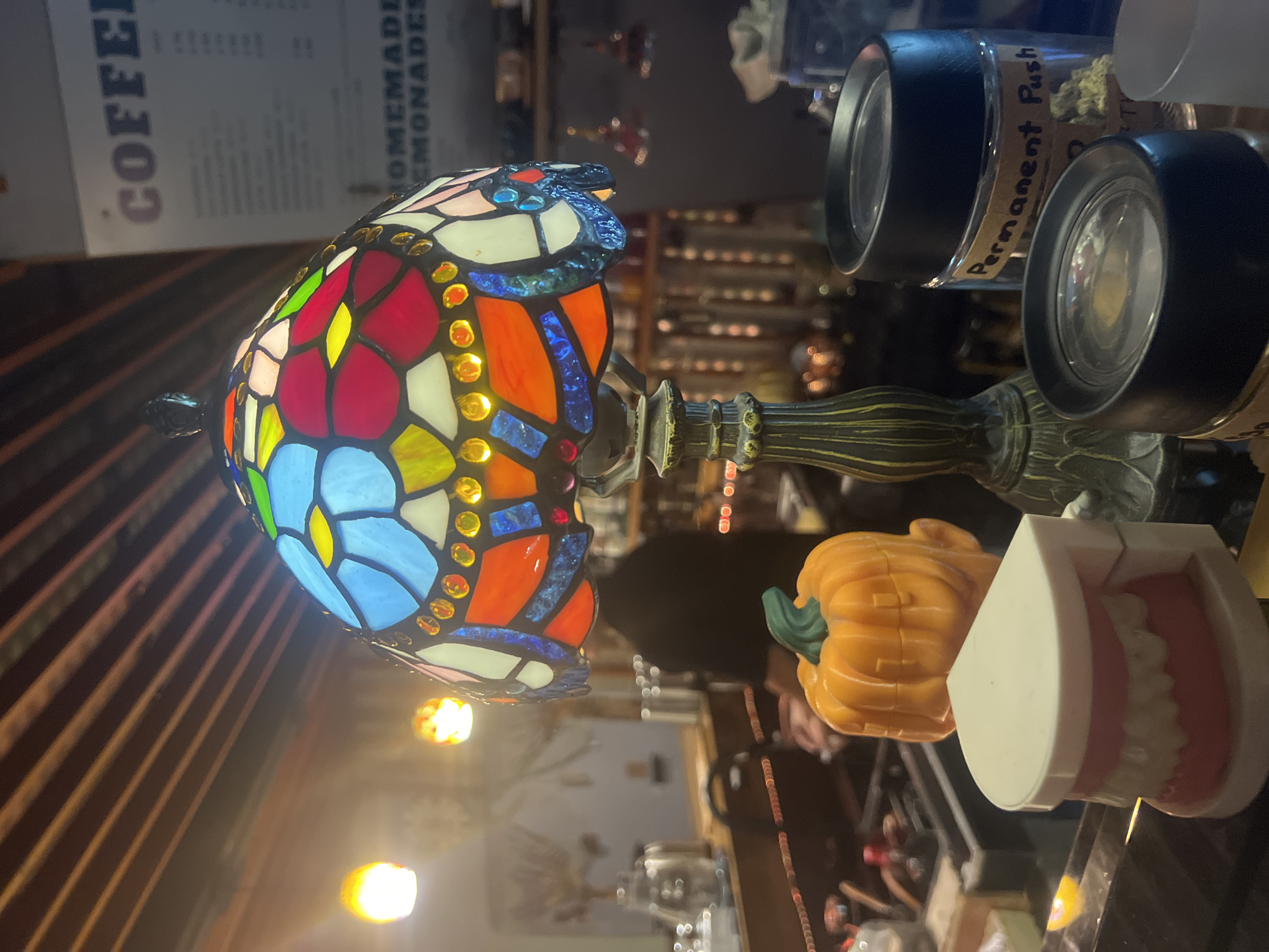 The image depicts a close-up view of a counter, likely in a cafe or shop. In the foreground, a set of dentures is displayed on the left, followed by a decorative orange pumpkin. Dominating the center is a Tiffany-style lamp with a colorful stained-glass shade, casting warm light. Behind the lamp, a person in a black shirt is visible, appearing to be working behind the counter. To the right, two glass jars with black lids are present; one is labeled "Permanent Push." In the background, a menu board with the word "COFFEE" at the top is partially visible. The environment appears to be indoors, with ambient lighting from overhead fixtures and the lamp. The setting is a commercial establishment in the city of Ko Samui District, Thailand.