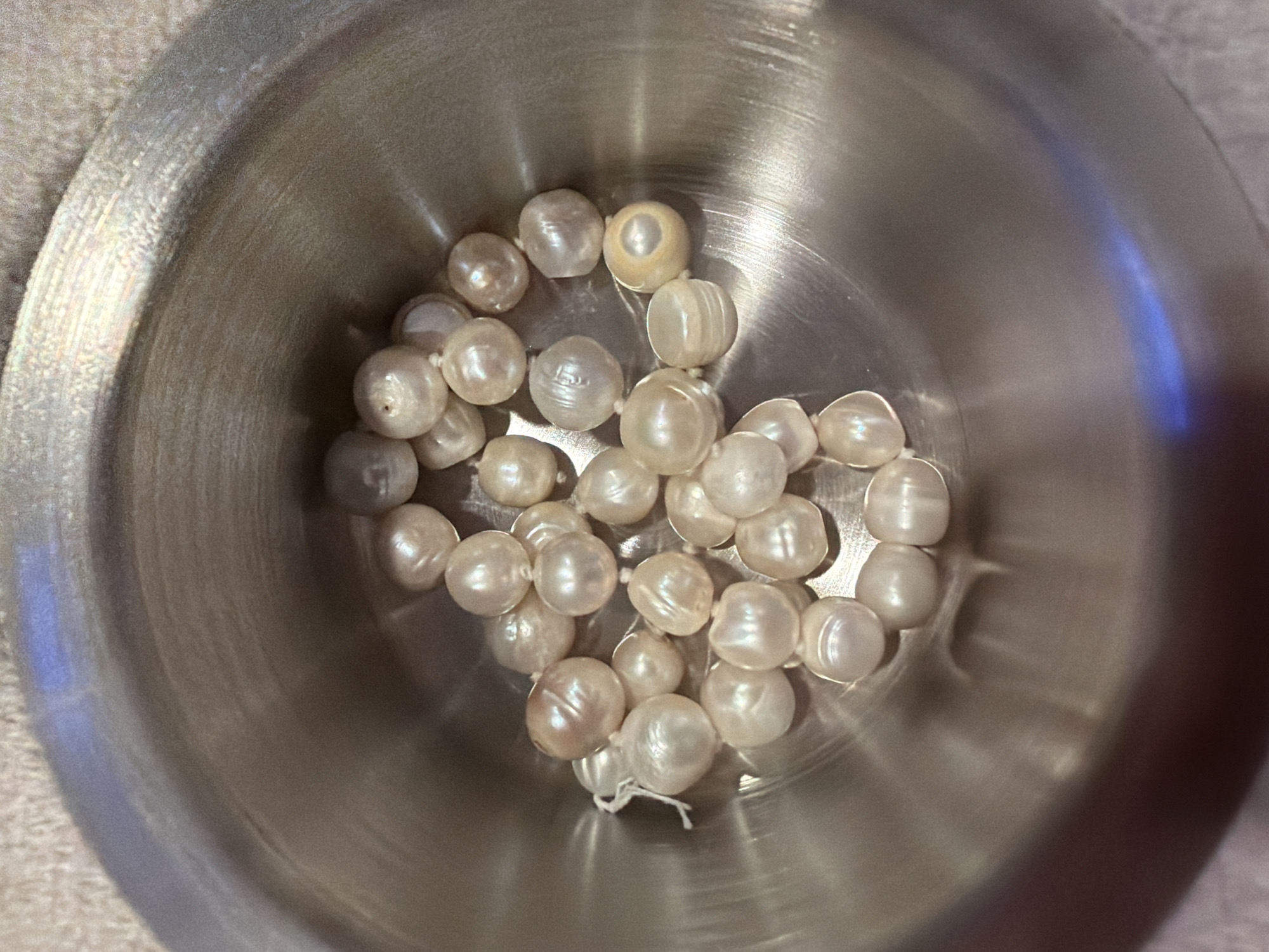 A top-down, close-up view displays approximately 30 to 40 irregularly shaped, baroque freshwater pearls resting in a polished stainless steel bowl. The pearls vary in size and exhibit off-white, cream, and faint pinkish-yellow hues with visible luster. Several pearls show prominent drill holes, and a segment of white string is visible among the pearls on the left side of the bowl. One pearl on the far right has a distinct yellow tone with a darker central spot. The reflective interior surface of the metal bowl shows numerous light scratches and strong glare spots, including an elongated blue reflection on the lower right interior. The bowl is situated on an out-of-focus, light beige, textured fabric surface. The scene is located in Bangkok, Thailand.