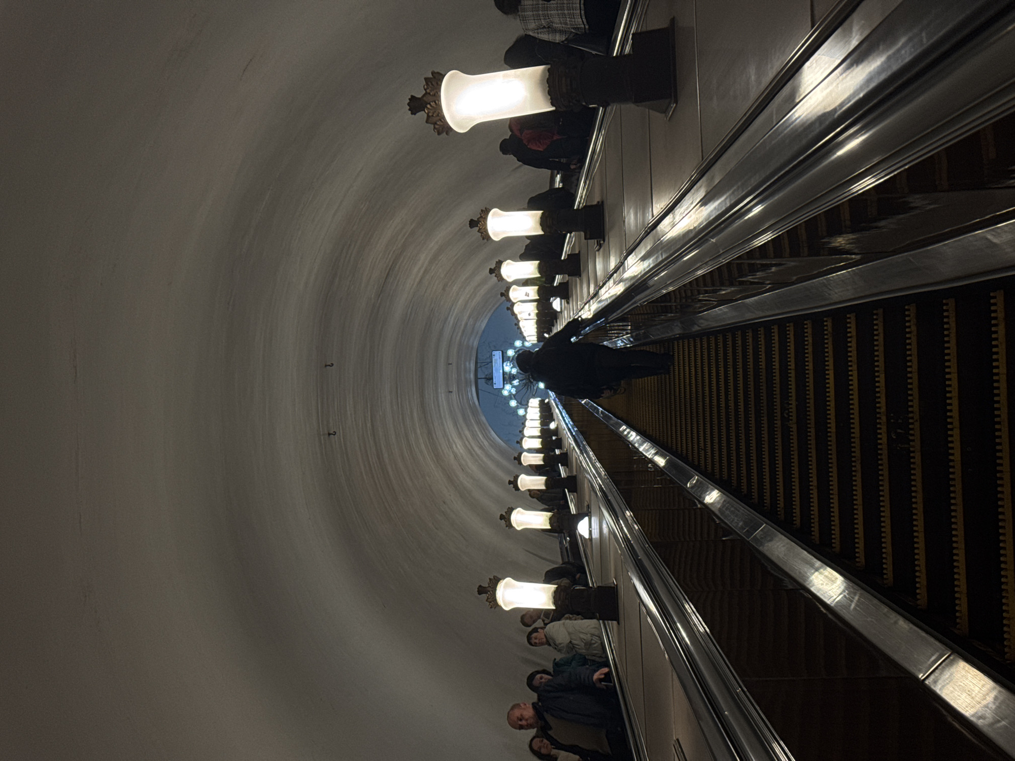 The image shows a long escalator going down into a subway station. People are standing on the escalator, going downward. The walls of the station are curved and lined with decorative lamps. The scene appears to be set in Moscow, Russia, most likely at the Park Pobedy metro station, known for its long escalators and distinctive architecture.