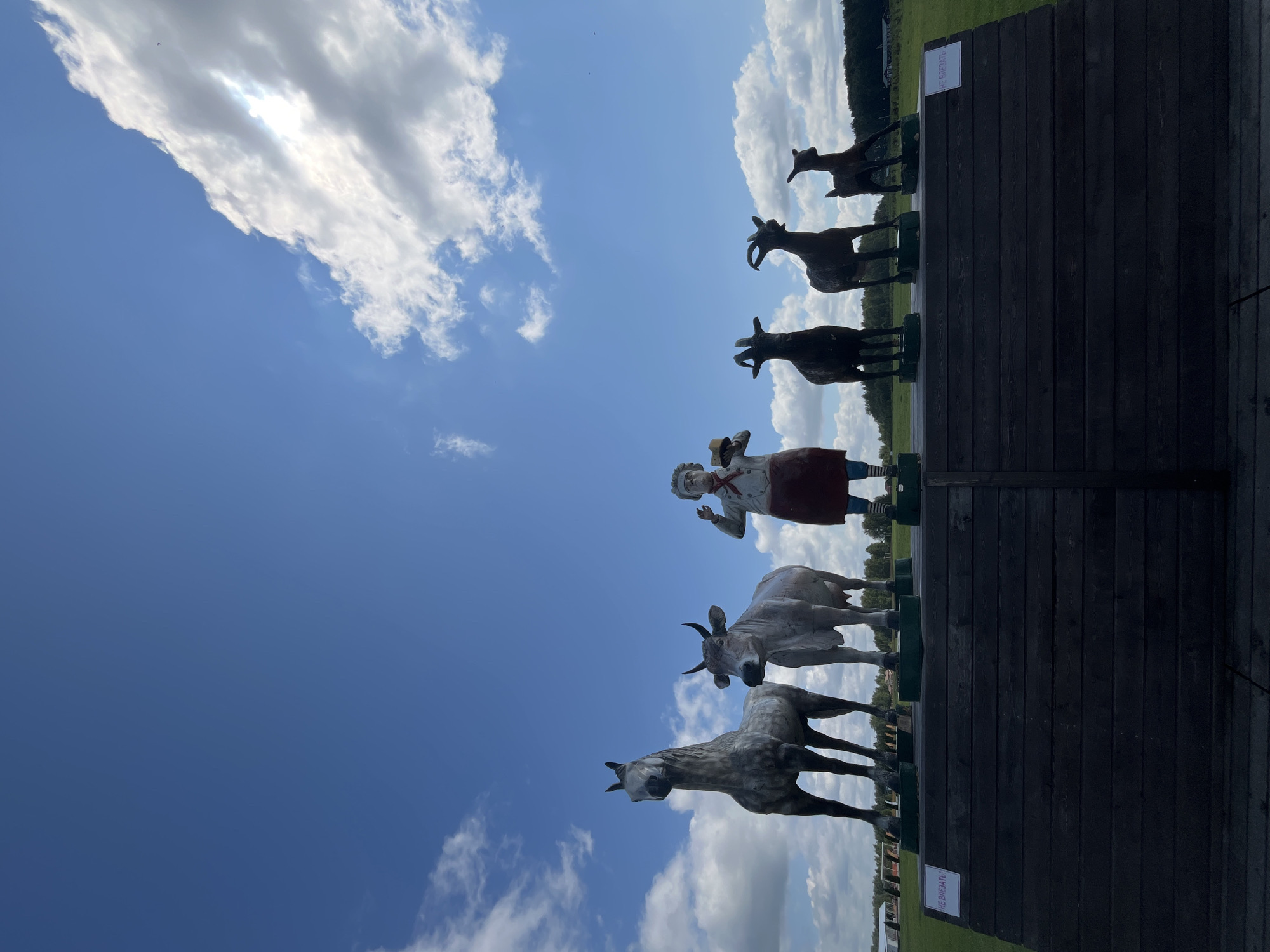 A low-angle outdoor shot shows a collection of statues of farm animals and a chef, standing on a dark wooden platform under a blue sky with scattered white clouds. The statues are arranged in a line, from left to right: a gray horse, a light brown cow, a chef in a red apron and white hat, a dark brown goat, another dark brown goat, and a small light brown goat. The chef statue is holding a light brown circular object, possibly a plate or a hat. The platform has two signs with red lettering; the left sign is partially visible and says "НЕ ВЛЕЗАЙТЕ!", meaning "Do not climb!" in Russian. The right sign is also partially visible with red lettering. Behind the platform, a grassy field stretches out, with trees and some structures in the distance. The location is identified as Akhlebaikha (Kovriginskii S/S), Russia. The lighting suggests it is daytime, with sunlight illuminating the scene.