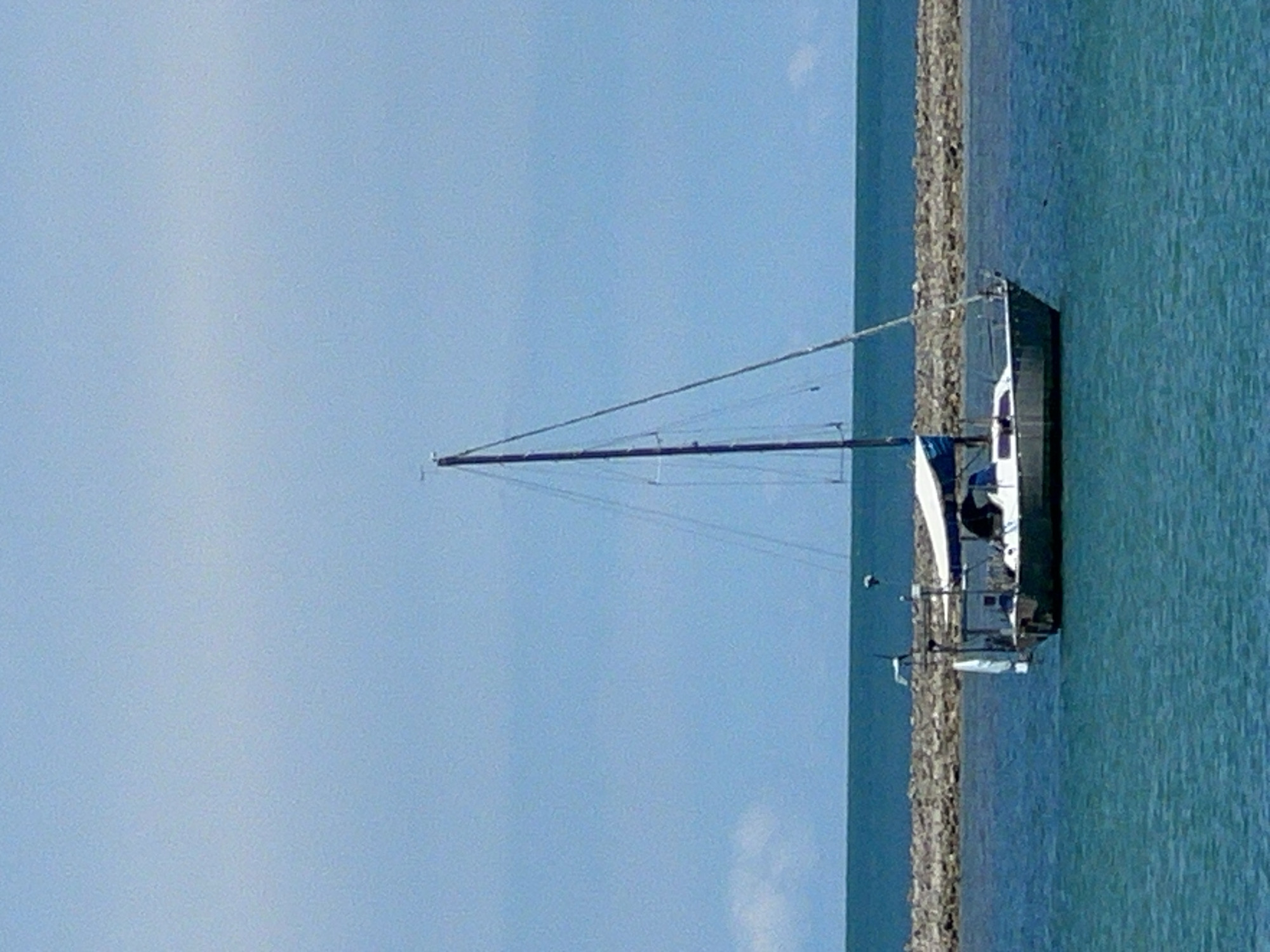 A sailboat with a dark hull and a white canopy is anchored in turquoise water. The sailboat has a tall mast with rigging and a blue sail partially furled. A person is visible inside the cockpit of the sailboat. In the background, a breakwater made of rough, gray stones extends horizontally across the frame, separating the turquoise water from a darker blue sea. The sky above is a clear, light blue with a few wispy white clouds on the horizon. This scene is located in Baan Hua Thanon, Thailand.