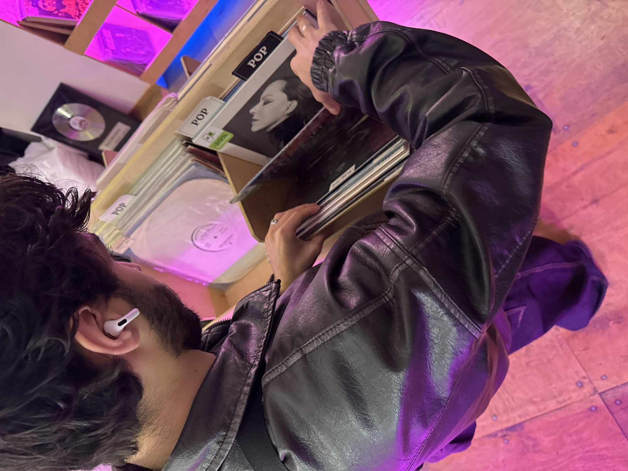 Here is a detailed description of the image:
A person wearing a black leather jacket, jeans, and a white wireless earbud is browsing through a bin of vinyl records. The records are labeled "POP". The environment appears to be a record store with pink and purple lighting. The city and country are Yerevan, Armenia. A person's hands are visible near the records, suggesting interaction with the merchandise. Also in view is an award plaque.
