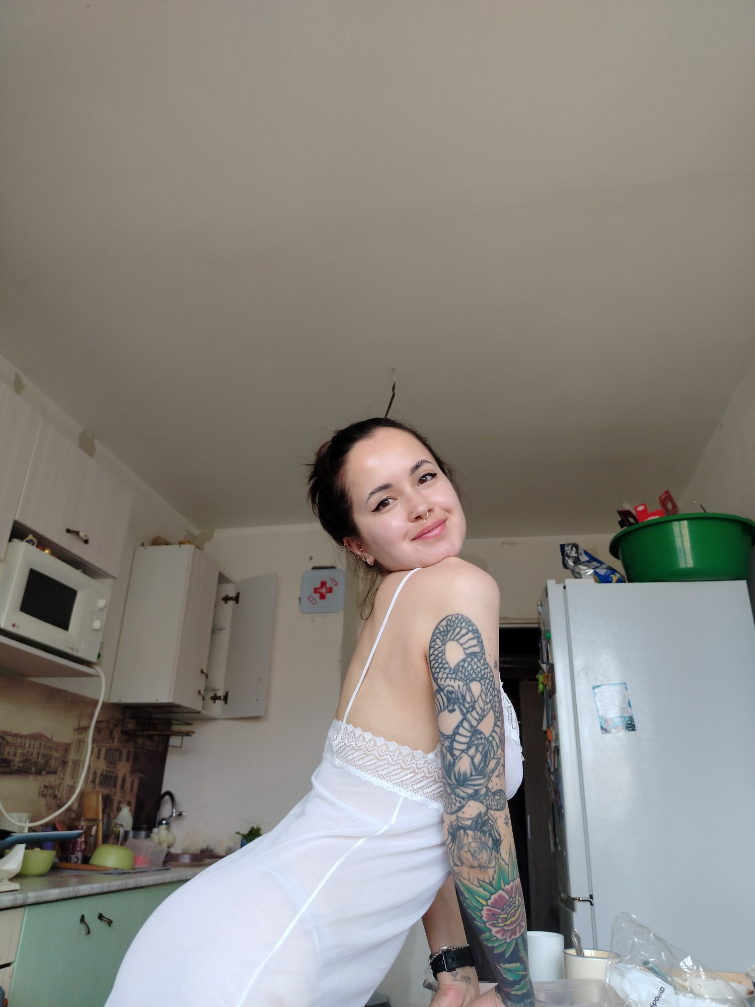 A young woman poses in a kitchen. She has dark hair pulled back, fair skin, and multiple piercings on her left ear and a nose ring. She is wearing a white, lace-trimmed nightgown. Her left arm is covered in a detailed tattoo featuring a snake and floral designs. The kitchen features white cabinets, a microwave, and a refrigerator. A green bowl sits atop the refrigerator. Other details include a watch on her left wrist, a landscape print backsplash, and a utility hook hanging from the ceiling. The location is Noviy, Russia.