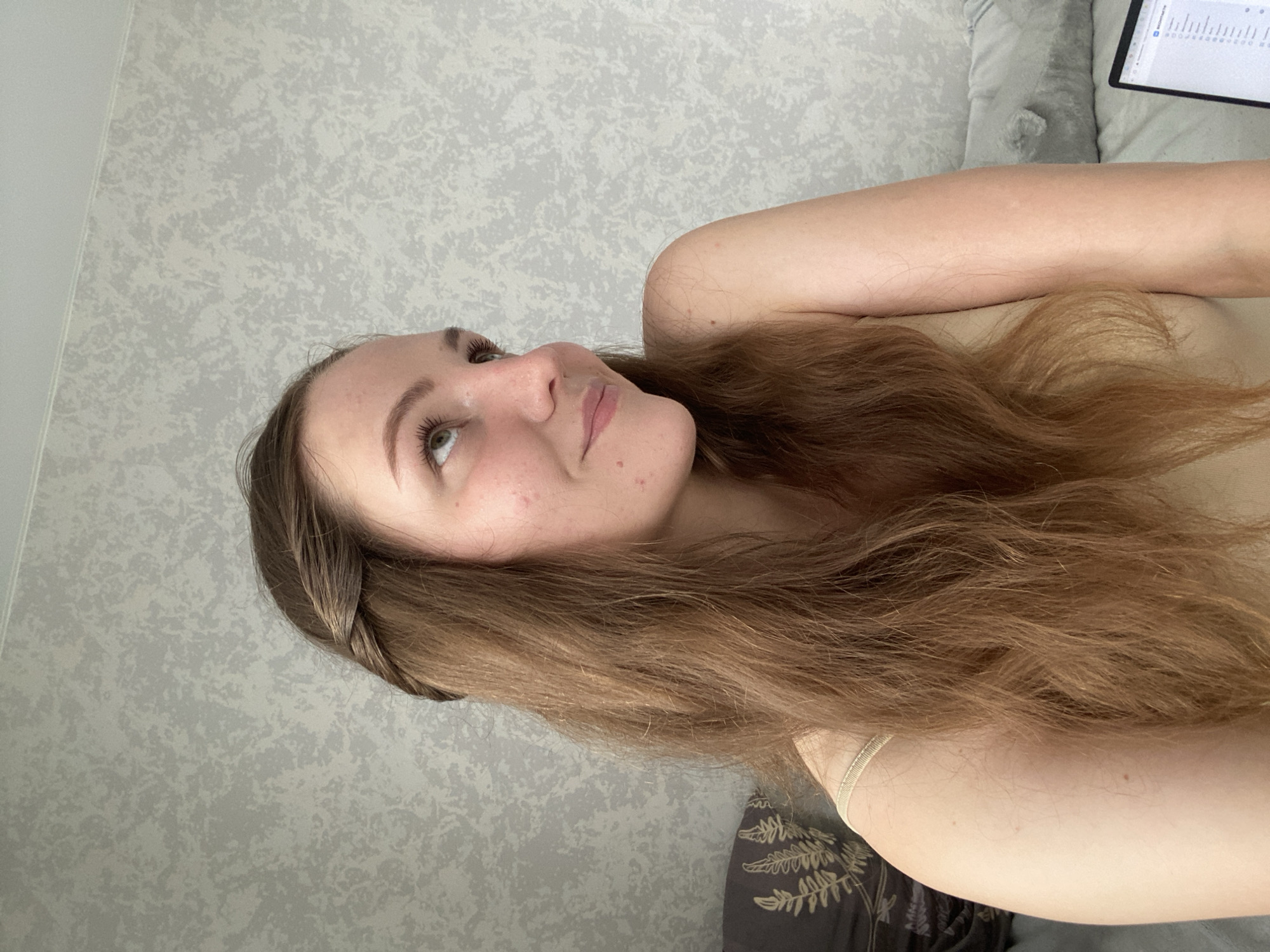 A young woman with long, brown, wavy hair is captured in a close-up, side-profile shot. A thin braid is visible on the left side of her head, accentuating her facial features. She is looking upwards and to her right, with a slight smile. Her skin is fair and shows some minor blemishes. She is wearing a light-colored tank top. The background consists of a textured, light gray wall with a subtle pattern, suggesting an indoor setting, possibly a bedroom, given the presence of pillows and a laptop visible on the right. The lighting is soft and natural, indicating daylight. No specific location in Izhevsk, Russia, can be determined from the image.