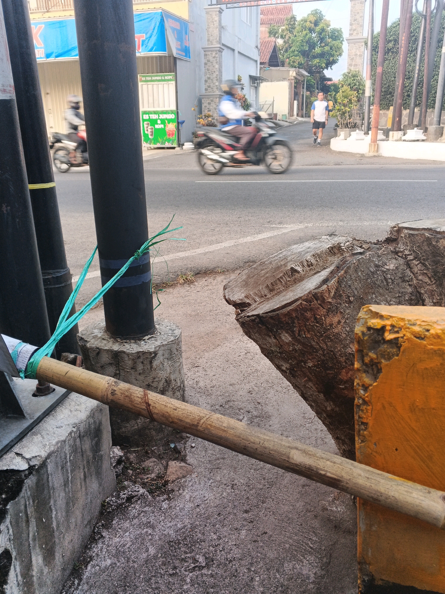 The image captures a street scene in Semarang, Indonesia, likely during the daytime. In the foreground, a large tree stump, roughly cut, dominates the right side of the frame, with a section of bright orange painted concrete next to it. A wooden pole, possibly part of a tool or barrier, lies horizontally across the lower portion of the image, secured to a concrete base by green twine. To the left, several dark, cylindrical poles stand vertically, one of which is also tied with the same green twine. The background reveals a street with two motorcycles. On the left, a motorcycle with a rider, blurred due to motion, is passing by. Further down the street, another motorcycle with a rider is visible. A man in a blue and white shirt and black shorts walks on the sidewalk on the right. Buildings line the street, including a shop with a blue banner and a green stand displaying "ES TEH JUMBO & KOPI" (Jumbo Iced Tea & Coffee). The environment appears to be an urban setting with a road, sidewalks, and buildings.