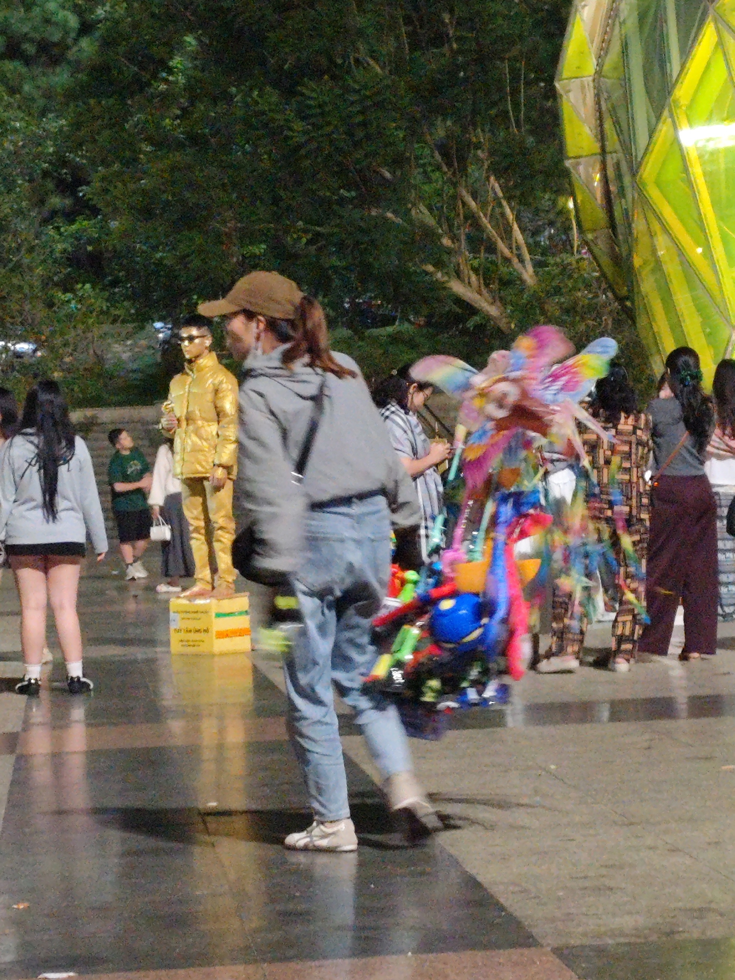 A street performer dressed in a gold outfit stands on a yellow box in Da Lat, Vietnam. Behind him, a person in a grey hoodie and blue jeans walks away from the camera, carrying a bundle of colorful, brightly lit balloon toys. Other people are visible in the background, including a woman in a patterned outfit and another in a grey shirt and dark pants. The environment appears to be an outdoor public space at night, with trees and a large, geometric, yellow structure to the right of the frame. The ground is wet, reflecting the lights.