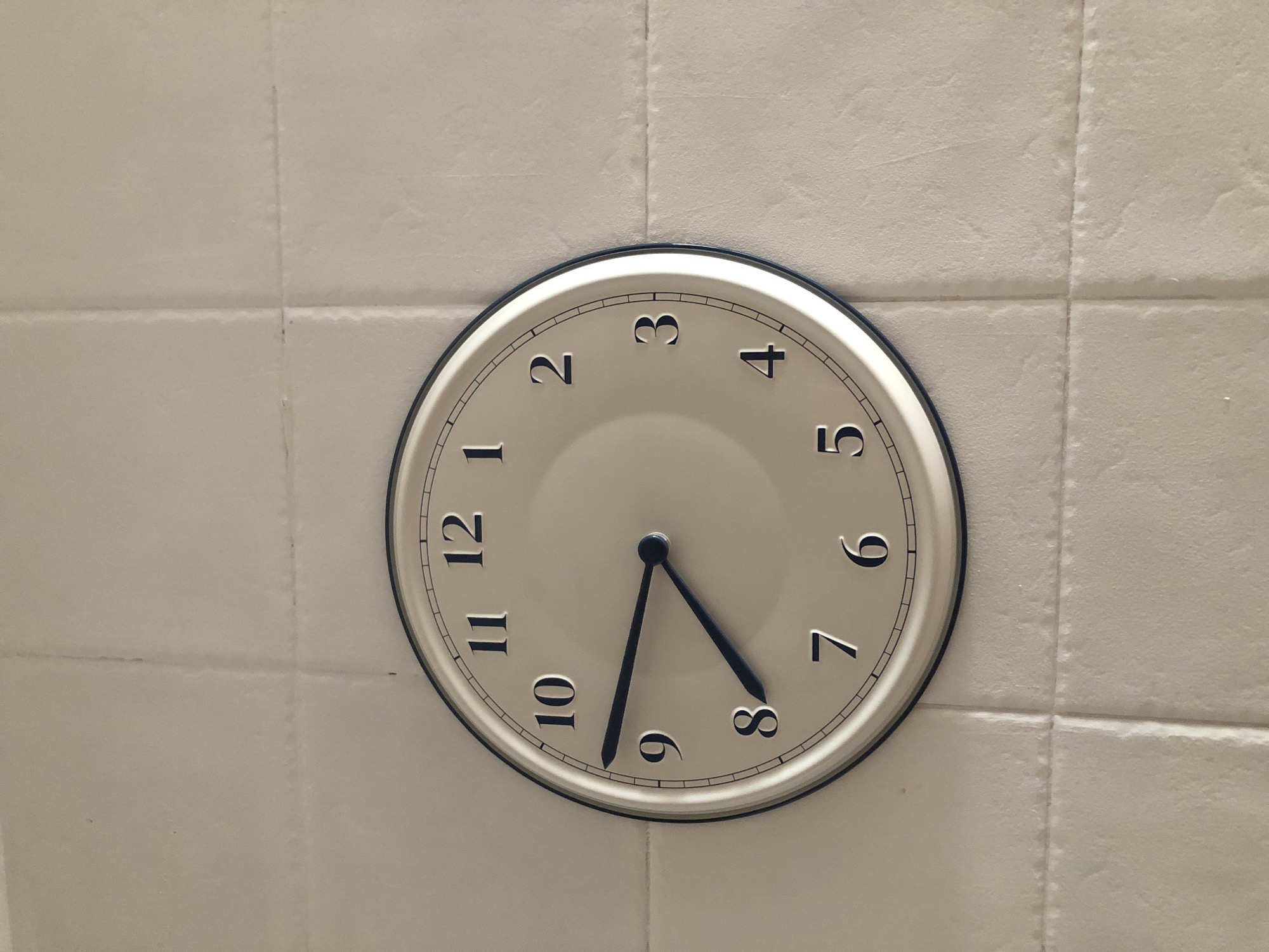 A close-up, high-angle shot shows a round analog clock mounted on a light-colored tiled wall. The clock face is cream-colored with black Arabic numerals and thin black hour markers. The clock has two black hands: an hour hand pointing slightly past the 8 and a minute hand pointing directly at the 9. The wall tiles are rectangular and arranged in a horizontal and vertical grid pattern. The image quality is good, with clear focus on the clock and the wall texture. The scene appears to be an indoor environment, possibly a kitchen or bathroom, based on the tiling. The specific location in Valencia, Spain, cannot be determined from the visual information provided in the image.