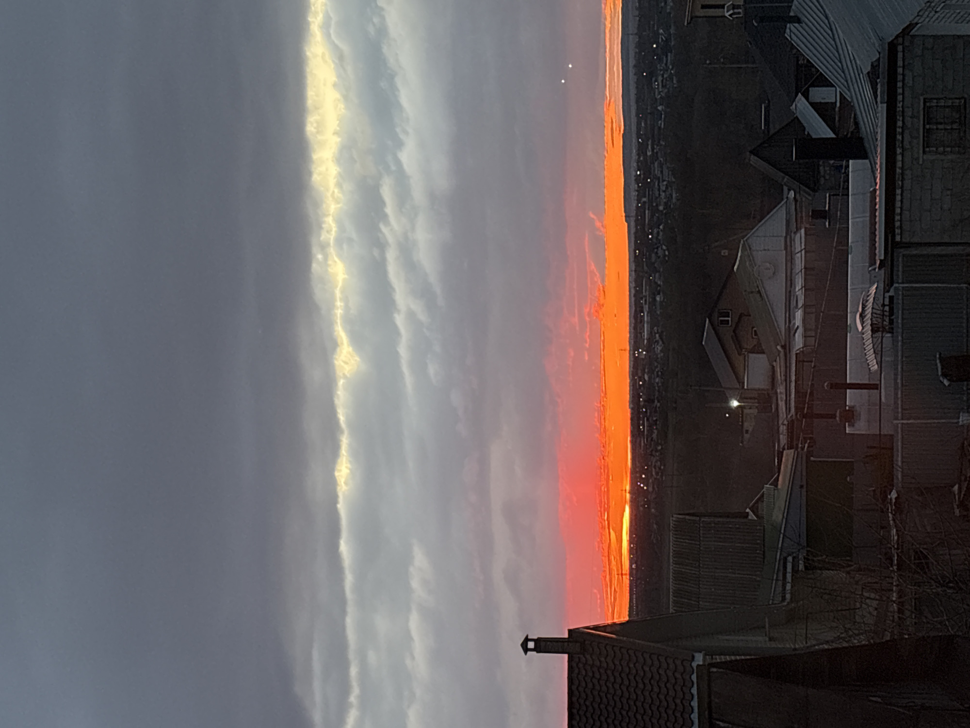 The image depicts a sunset in Ulyanovsk, Russia, viewed from an elevated position, possibly a rooftop. The sky transitions from a deep gray on the left to a vibrant orange and red on the right horizon, suggesting the sun is setting. A band of light-colored clouds runs vertically through the center. Buildings and structures are visible in the foreground, their details obscured by shadow. Lights are visible along the horizon indicating the city.