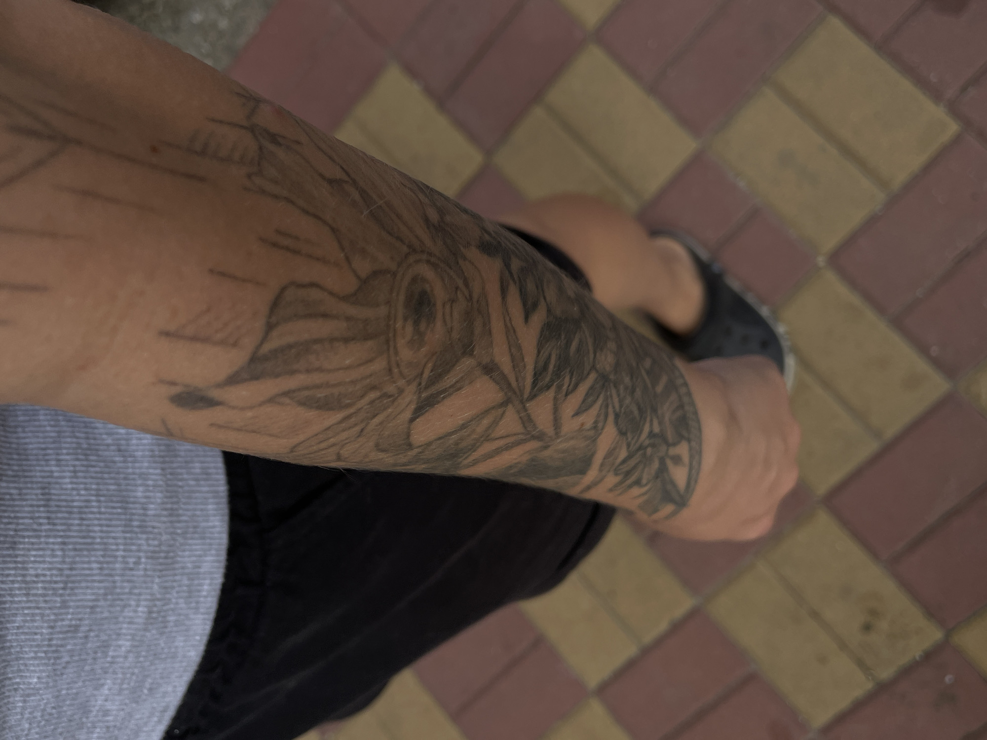 The image shows a person's tattooed arm, with the tattoo depicting a sunflower and possibly leaves. The person is wearing a grey ribbed tank top and black shorts, with black slip-on shoes visible on their foot. They are standing on a pathway paved with reddish-brown and yellow rectangular tiles. The background is out of focus, but suggests an outdoor environment. There are no discernible landmarks or indicators of the specific location within Rostov-on-Don, Russia. The lighting suggests it is daytime. The image is taken from a high angle, looking down at the person's arm and legs.