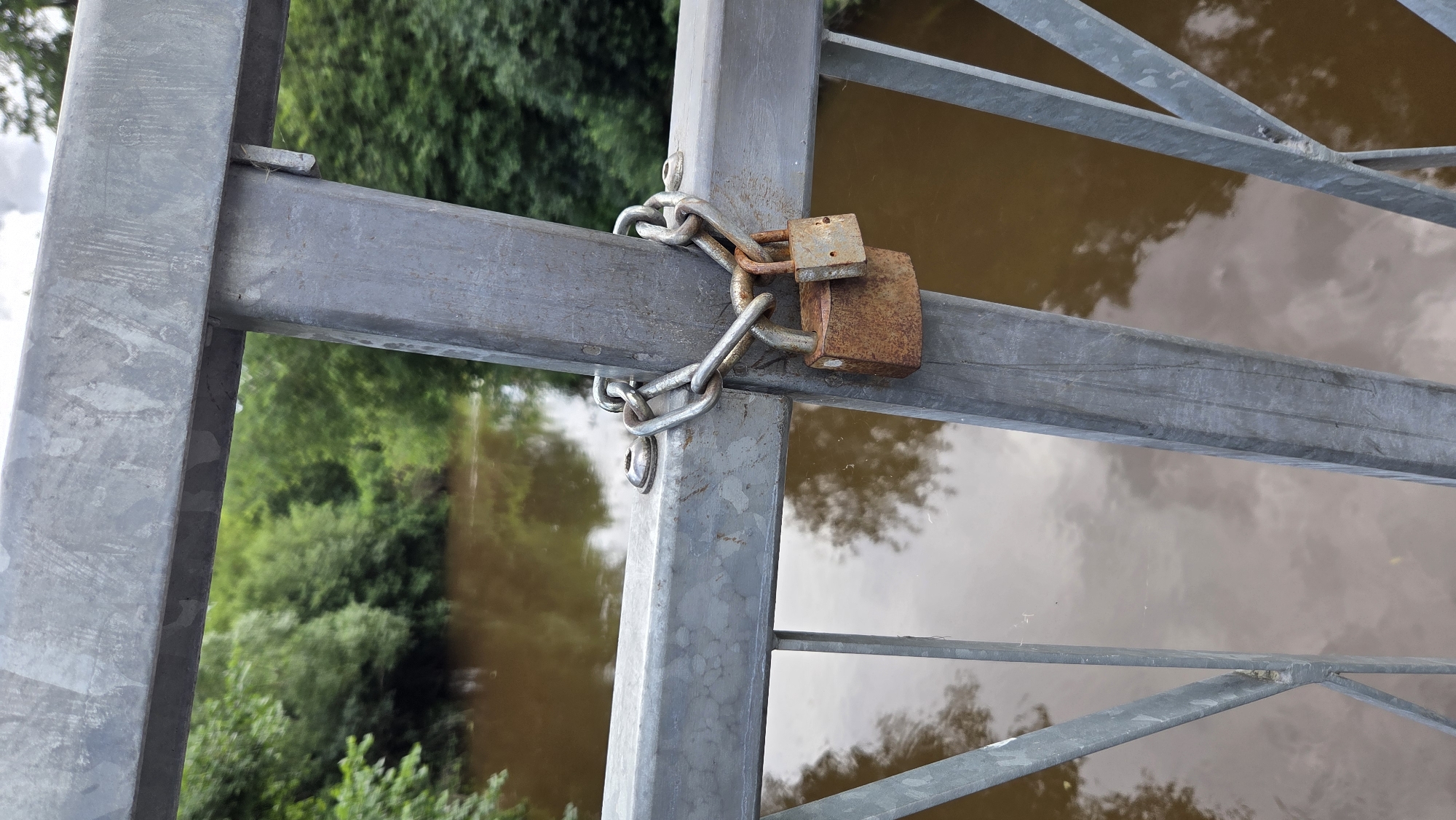 The image shows a close-up view of a metal gate with two rusty padlocks securing it with a chain. The gate appears to be part of a bridge or a fence overlooking a body of water, possibly a river or canal, with murky brown water reflecting the sky and surrounding greenery. The background is filled with lush green trees, suggesting an outdoor natural setting.
The padlocks are made of metal and have a weathered, rusty appearance, indicating they have been exposed to the elements for some time. The chain linking them is also metallic and appears to be of a robust design. The gate itself is constructed from thick metal beams and bars, painted in a dull gray, and shows signs of wear and tear. One of the gate's vertical support beams has a small, flat piece of metal inserted into a gap, possibly a shim or a repair.
The scene is brightly lit, suggesting it was taken during daylight hours. The sky's reflection on the water indicates some cloud cover. There is no visible text in the image. The overall impression is one of security and perhaps a sense of being closed off or restricted, given the locked gate. The location context of Ūlupji, Latvia, places this scene in a rural or semi-rural area of Latvia.