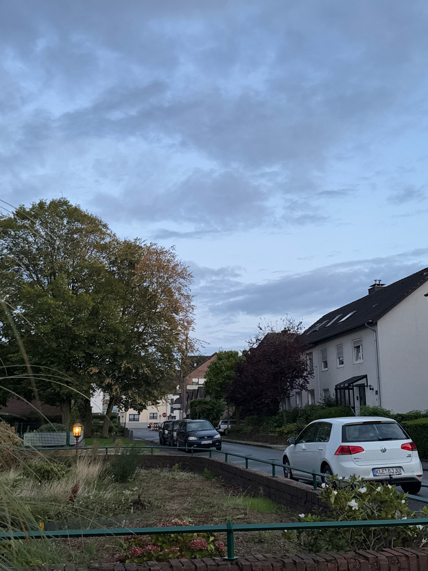 The image depicts a residential street in the city of Cleves, Germany. The scene is set under a cloudy sky, with a mix of green and autumnal foliage visible on trees lining the street. Several cars are parked and in motion. A white Volkswagen Golf is the most prominent vehicle, with a German license plate reading "KLE-BJ 30". Adjacent to it, a darker colored car, possibly an SUV, is also visible. Residential buildings with dark roofs and white facades line the right side of the street. In the foreground, there is a low brick wall and vegetation, including tall grasses and some flowering plants. A street lamp with an illuminated amber light is present on the left side of the image. The overall impression is a calm, suburban street scene, likely captured in the late afternoon or early evening.